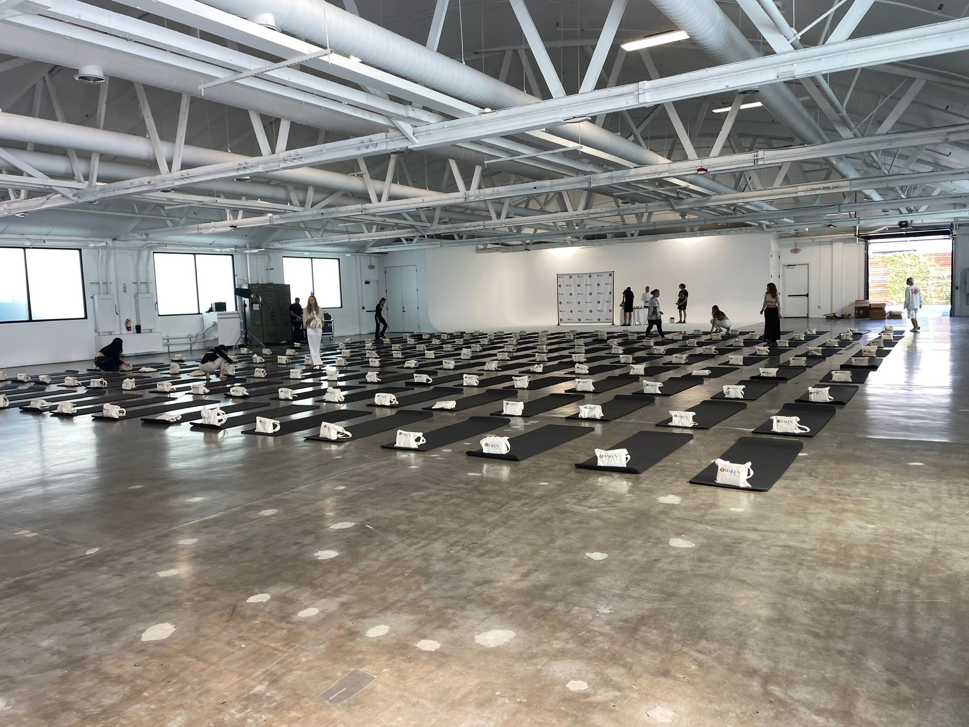 An empty, spacious room set up for a yoga or fitness class with rows of black mats on a light gray concrete floor.
