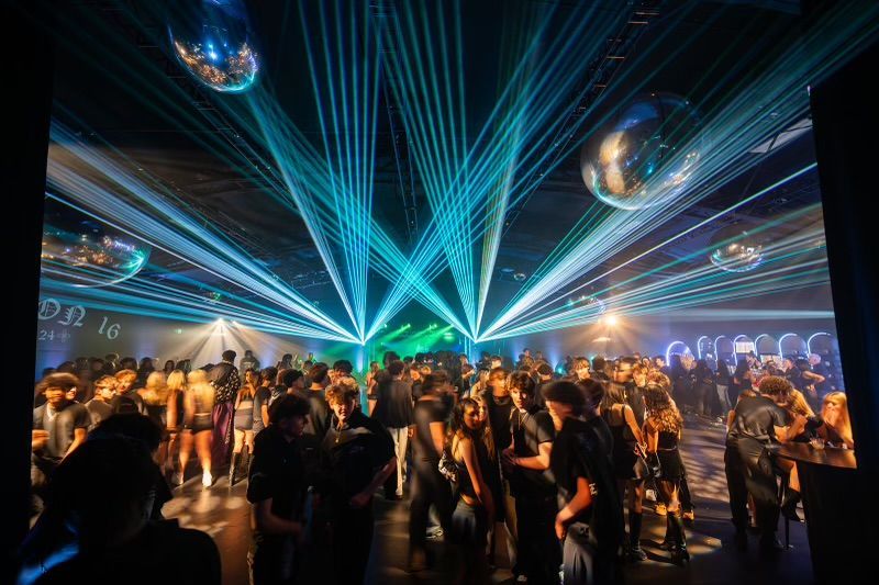 A crowded nightclub interior with people dancing under bright blue laser beams and suspended disco balls.