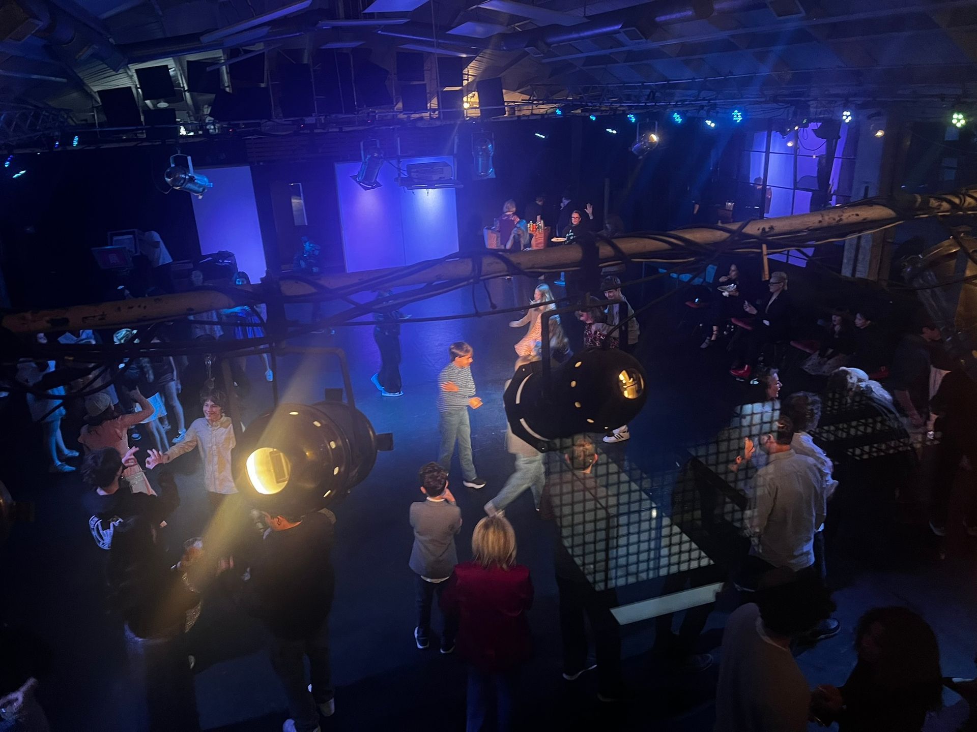 A dimly lit event space with stage lights, people gathering around display cases, and blue ambient lighting.