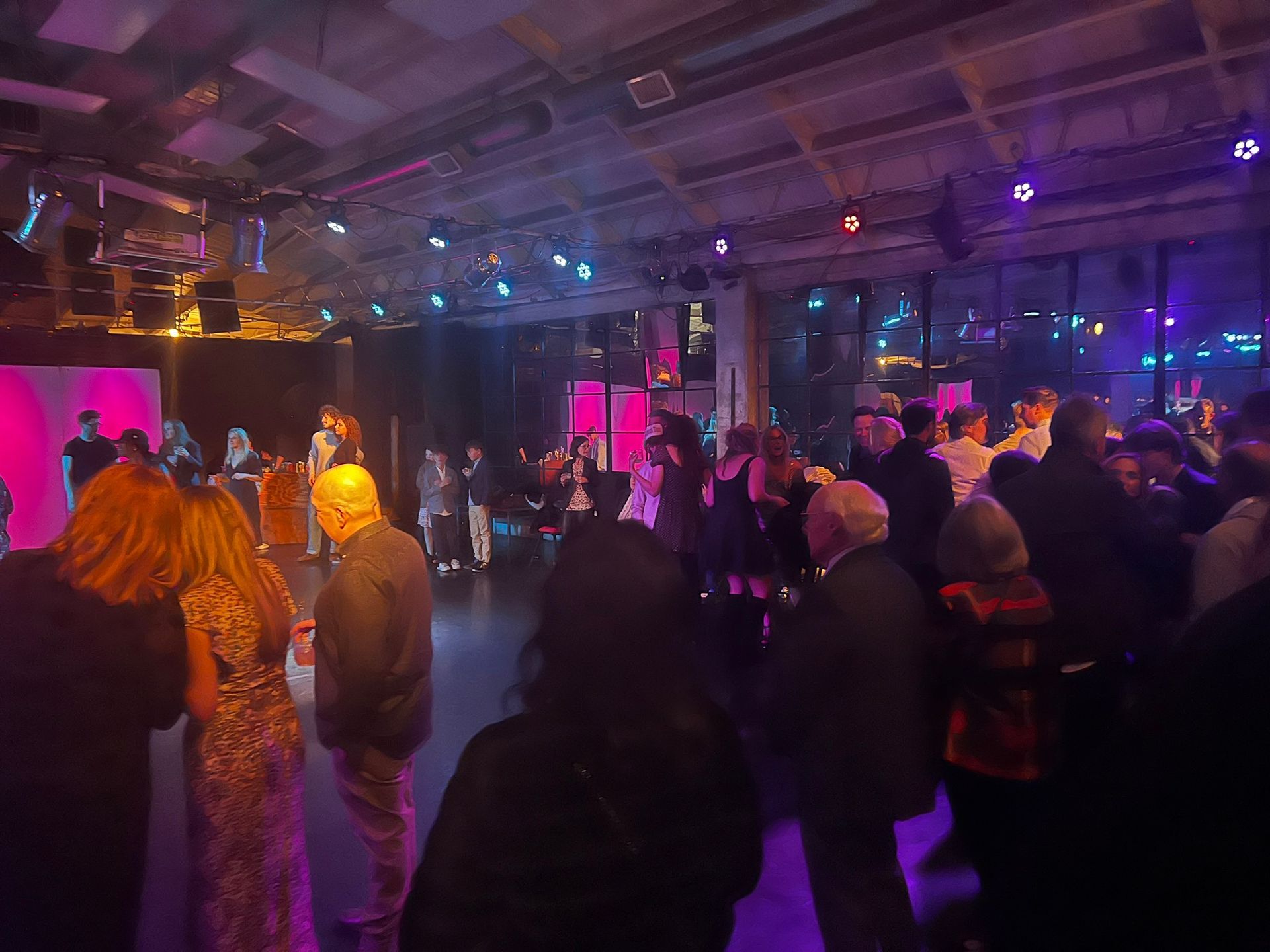 People gather in a dim, purple-lit indoor space with industrial ceilings and stage lights, appearing to be at an event.