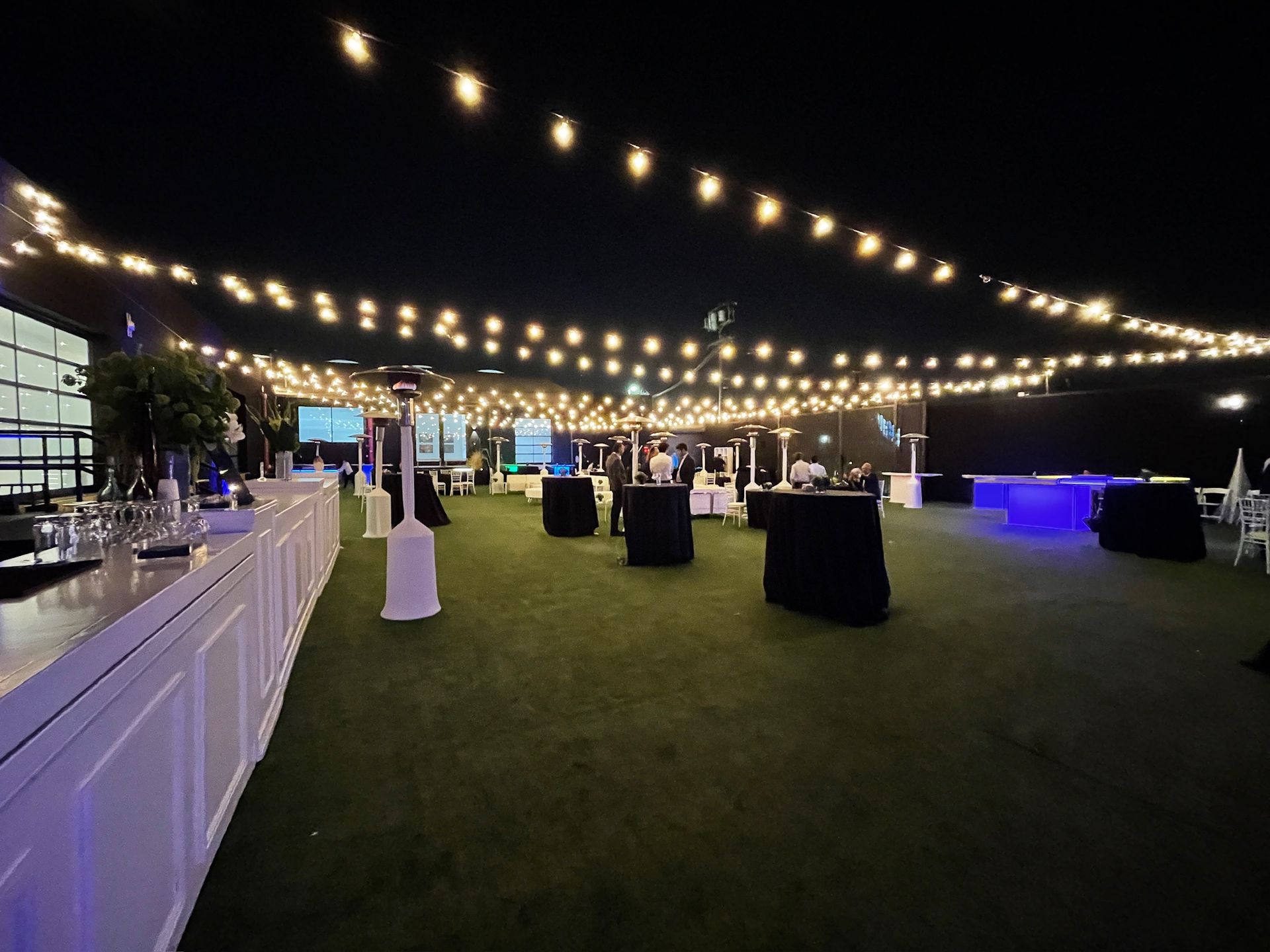 An outdoor evening event space with artificial grass, black-clothed cocktail tables, and glowing string lights overhead.