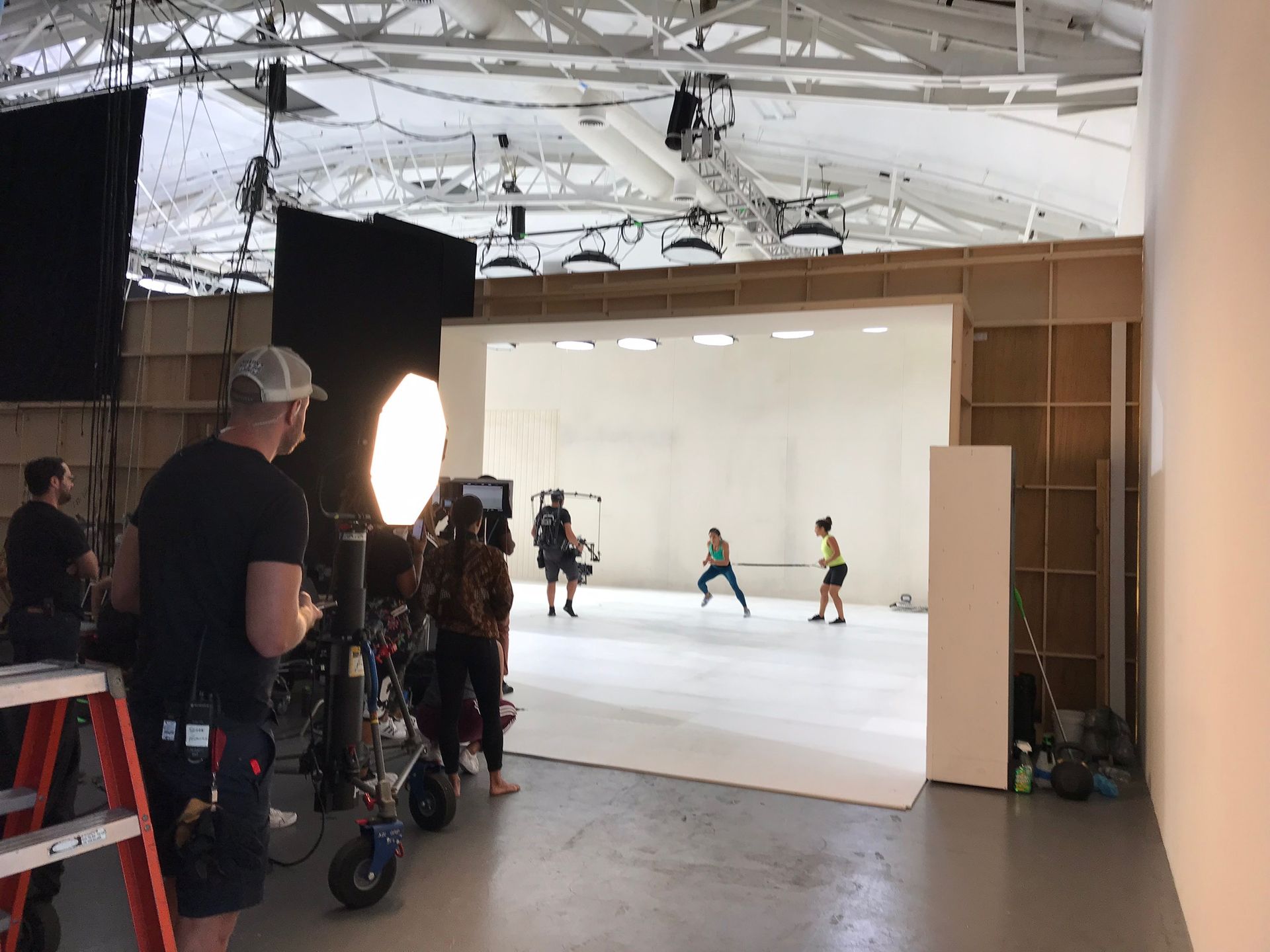 A film crew records two people moving across a brightly lit, white studio stage with professional equipment and lighting.