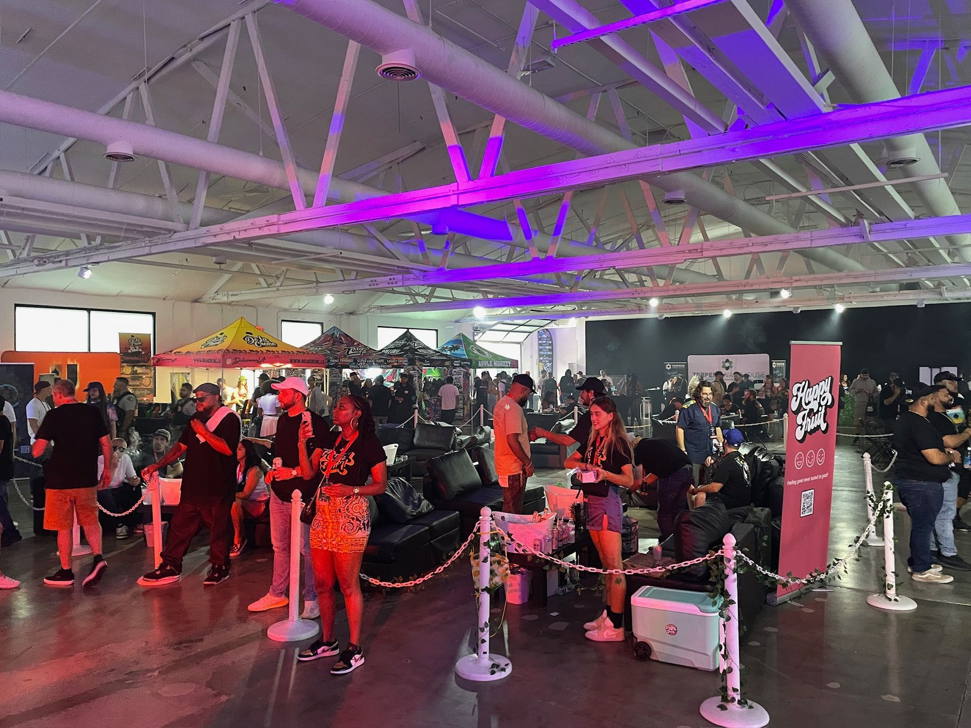 People stand in line at a trade show exhibit hall under purple lighting, with booths and equipment visible in the space.