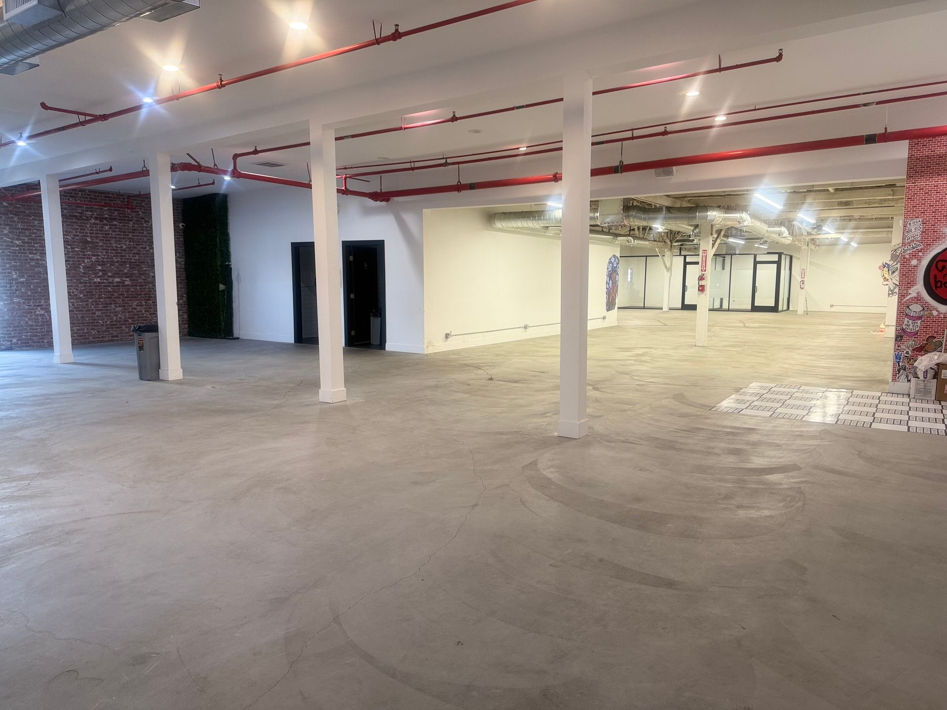 A spacious, empty industrial-style room with concrete floors, white pillars, and exposed red ceiling pipes.
