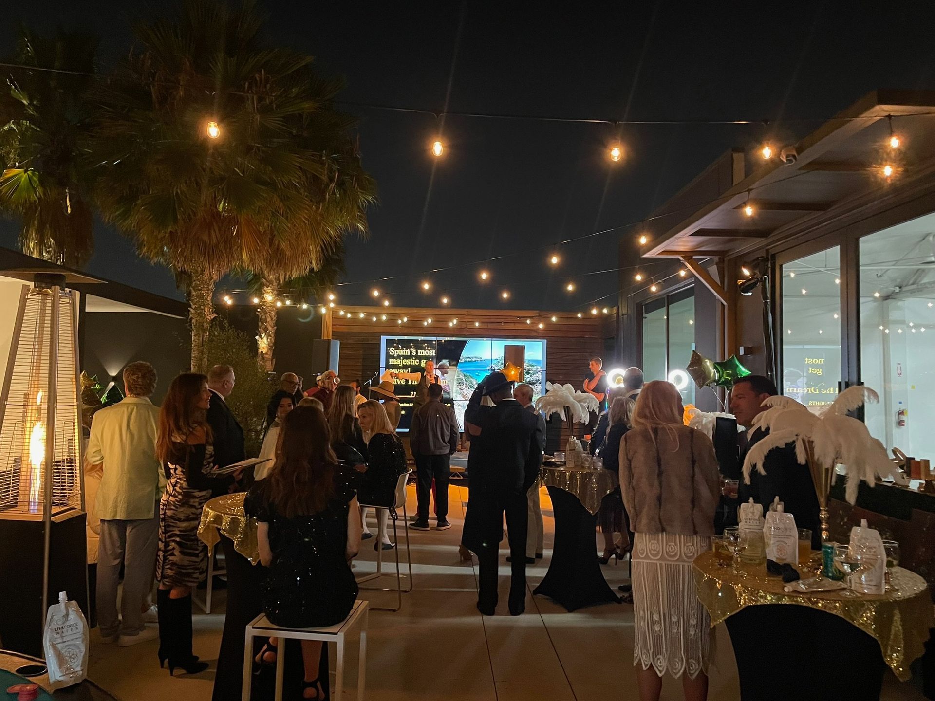 People socialize at an outdoor evening event decorated with string lights, palm trees, and tables with gold accents.