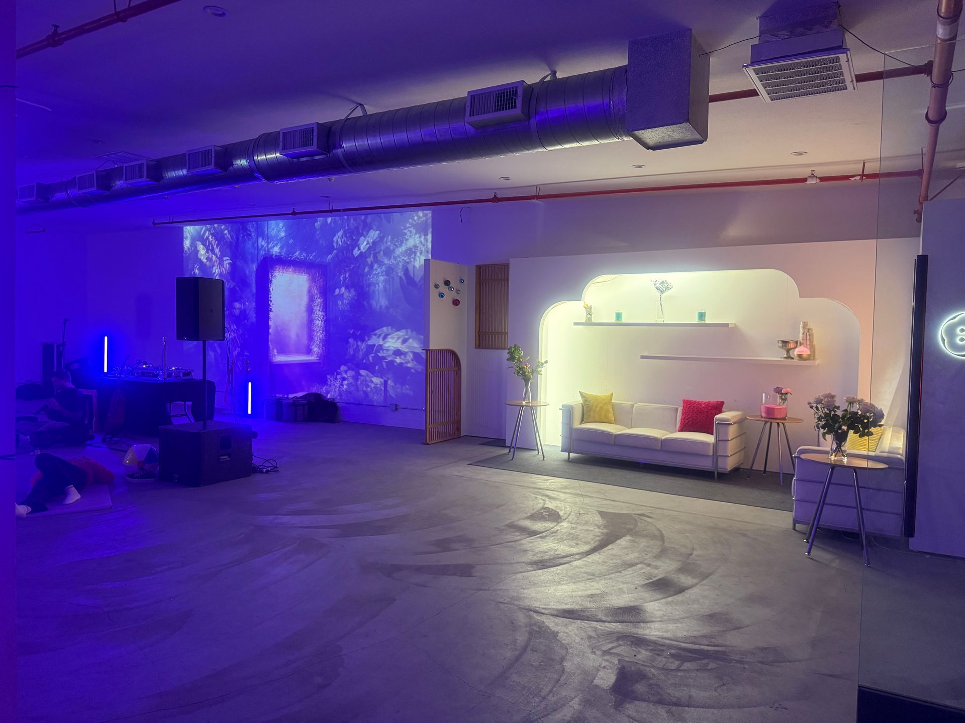 A dimly lit event space with a projection of a forest on a wall, a white sofa, and industrial-style exposed ceiling pipes.