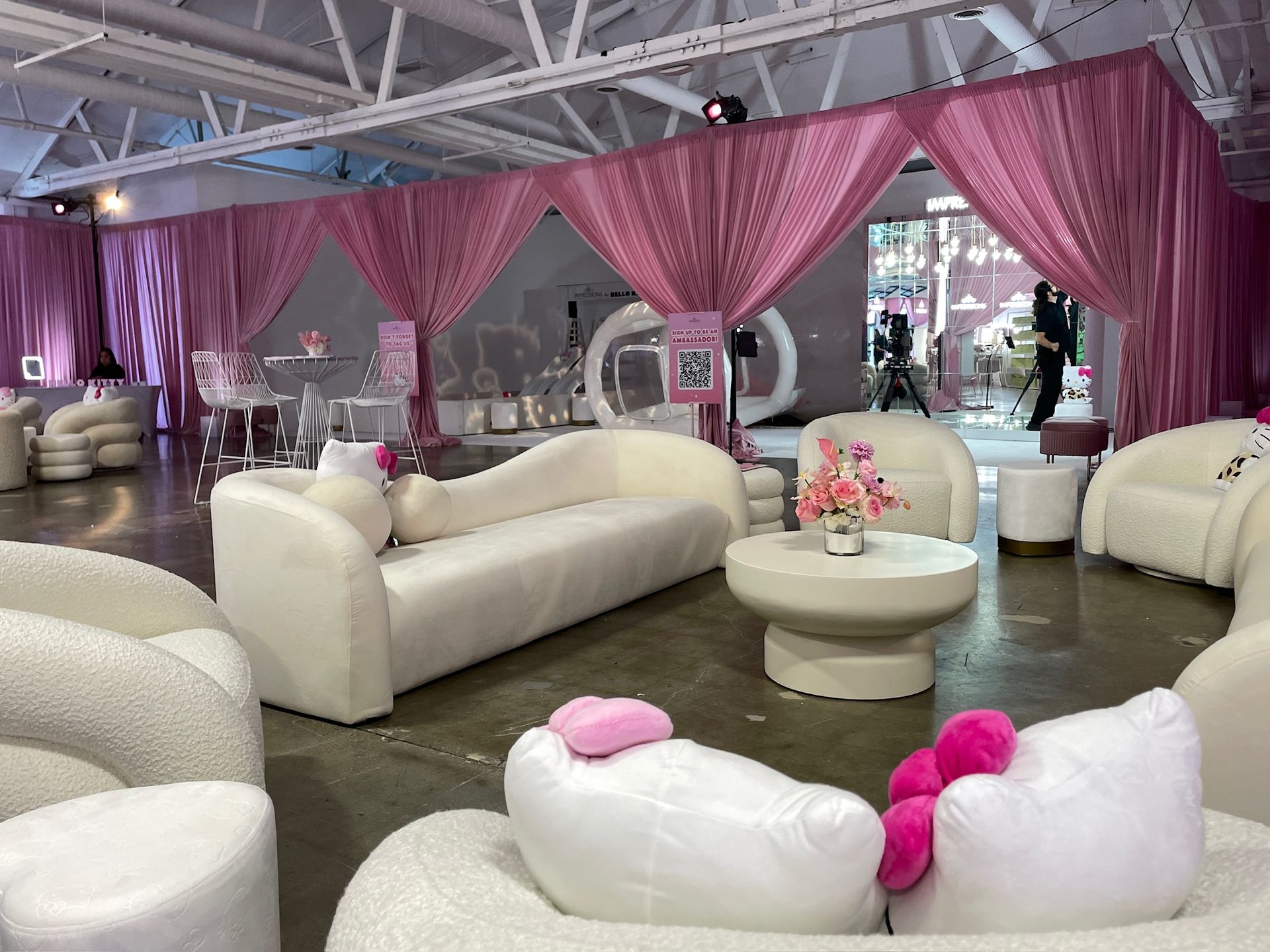 A lounge with white curved furniture and pink curtains, featuring Hello Kitty-themed pillows in the foreground.