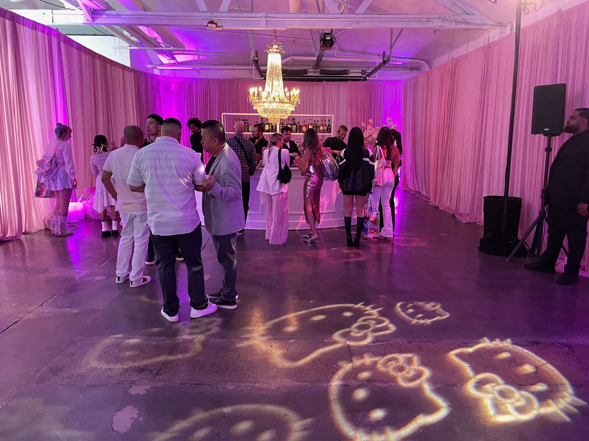 A brightly lit room decorated in pink, with guests mingling near a bar and Hello Kitty projections on the floor.