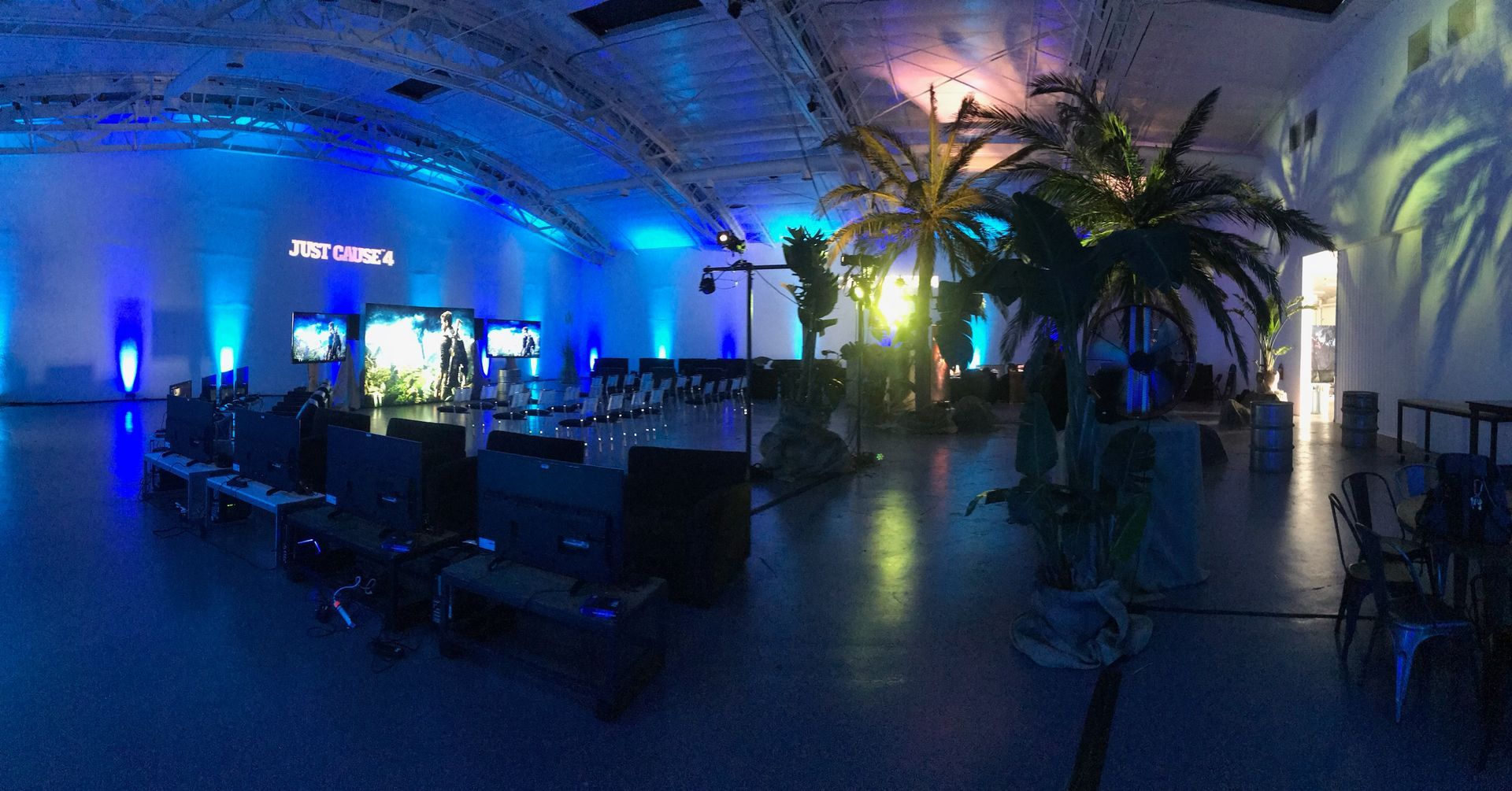 An event space with blue accent lighting, indoor palm trees, and gaming screens set up on tables.