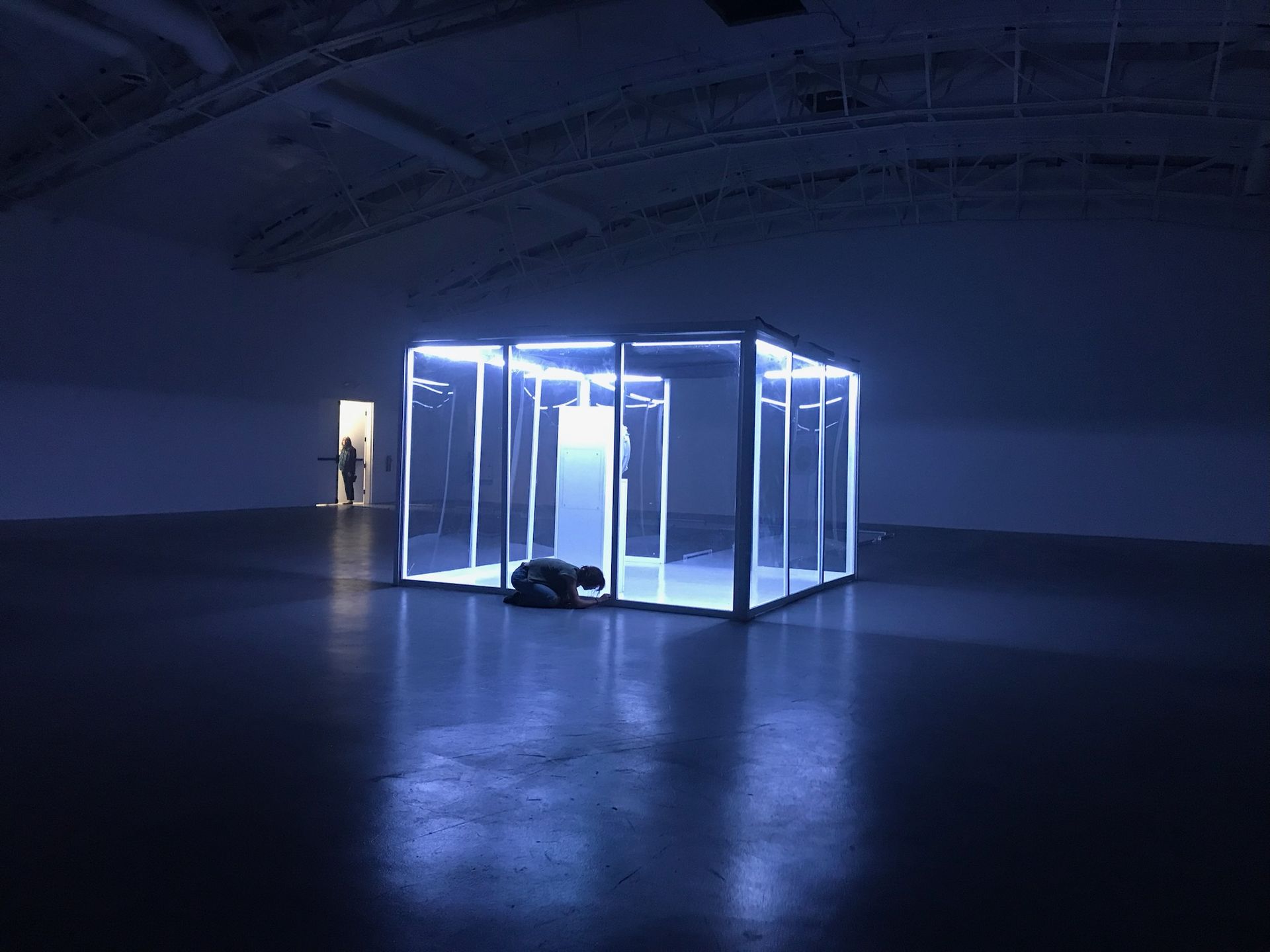 A person crouches inside a glowing, rectangular glass installation in a dimly lit, vast art gallery space.
