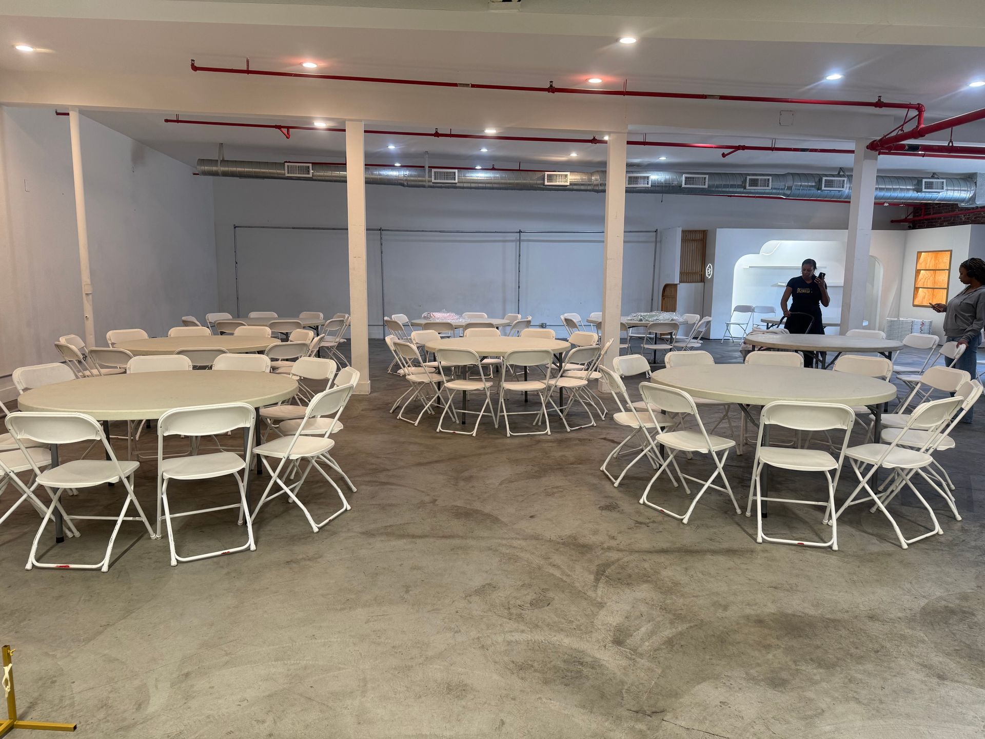 A large, open event space with white walls, exposed red ceiling pipes, and round tables set with white folding chairs.
