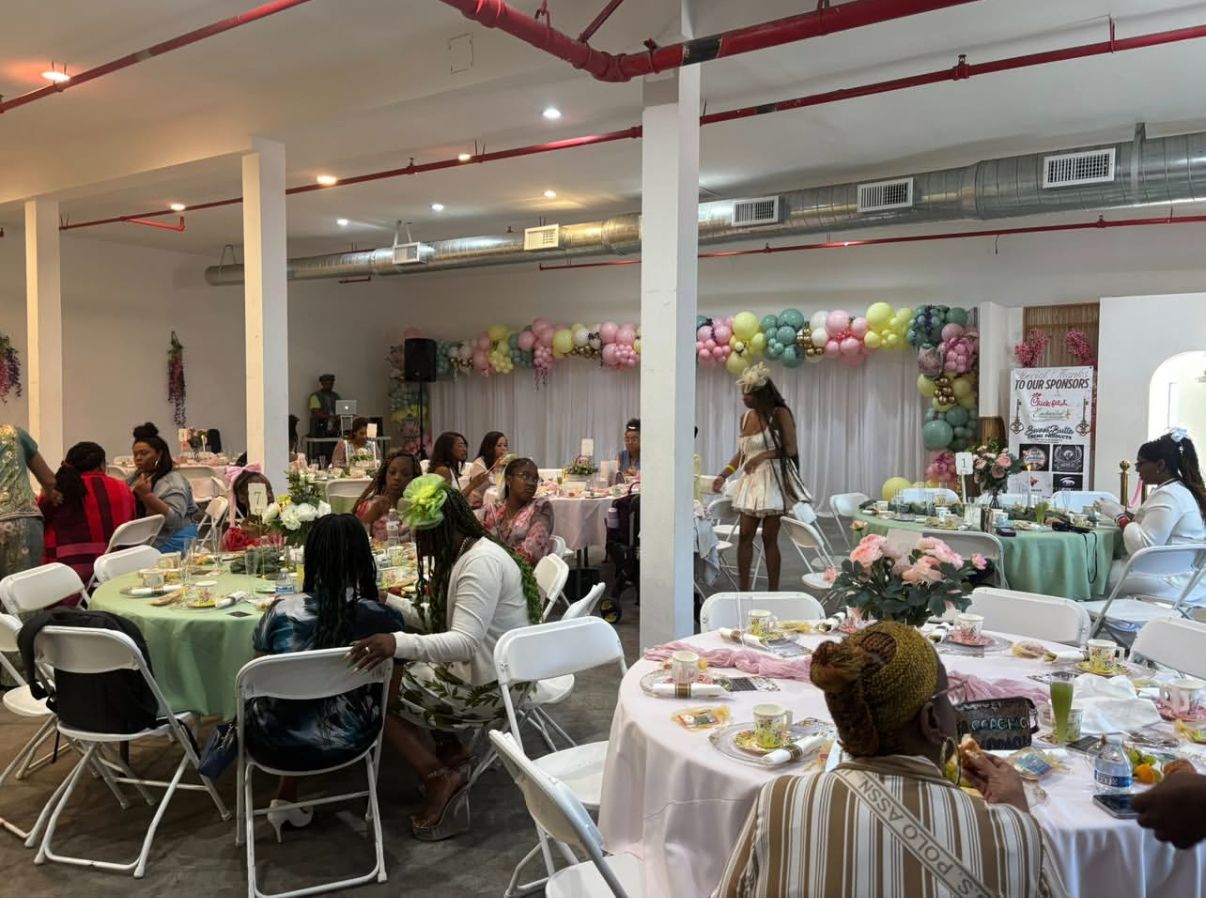 People attend an event in a hall decorated with pastel balloons, sitting at round tables with light green and white linens.
