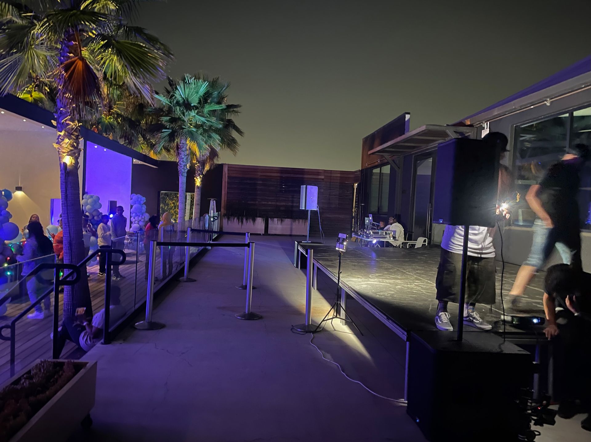 People gather on an outdoor rooftop terrace at night, featuring a stage with audio equipment, palm trees, and string lights.