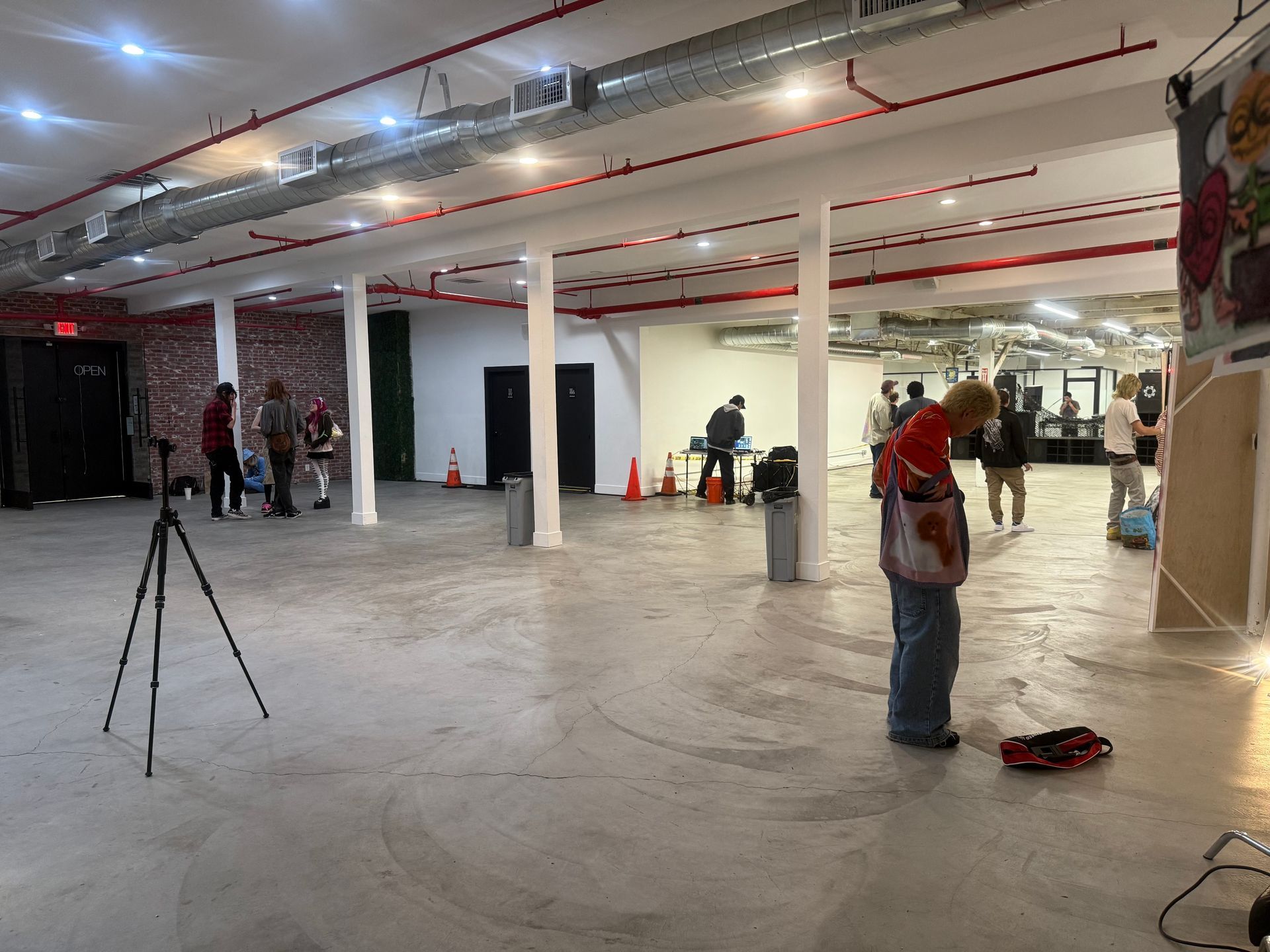 People set up equipment in a large, open industrial space with white pillars, concrete floors, and exposed red piping.