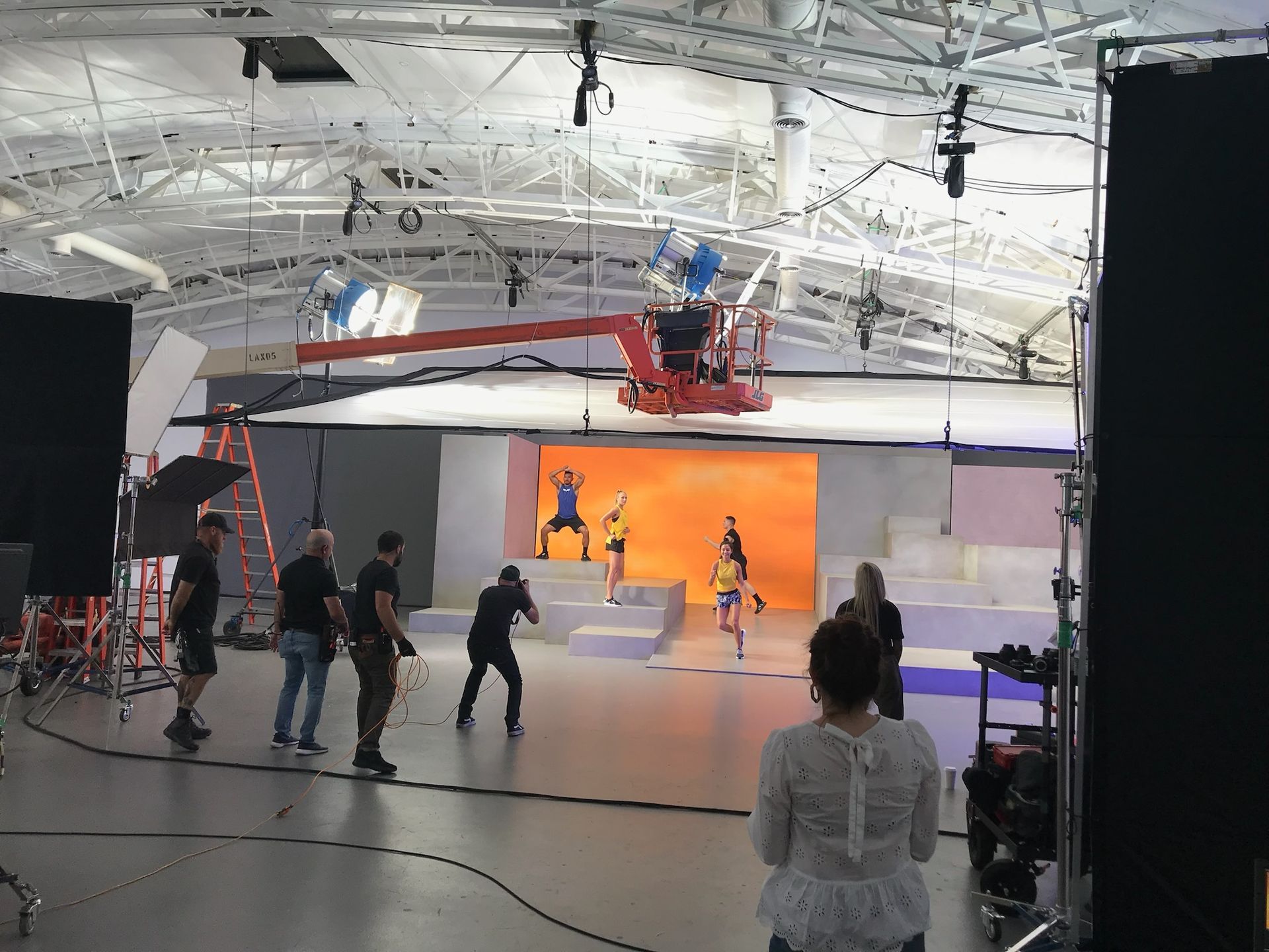 A film crew works in a studio with a bright orange backdrop and raised white platforms where performers are posing.