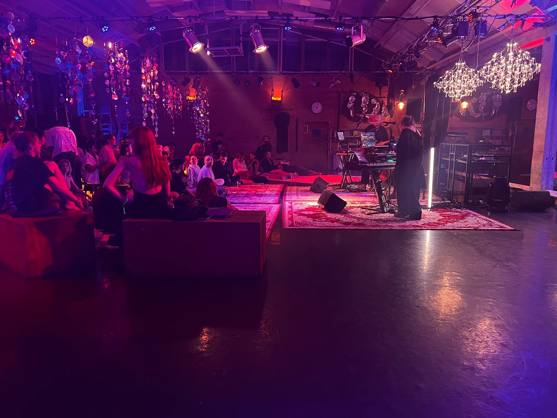 A person plays a keyboard at a concert in a dimly lit venue with red lighting, rugs, and sparkling chandeliers.