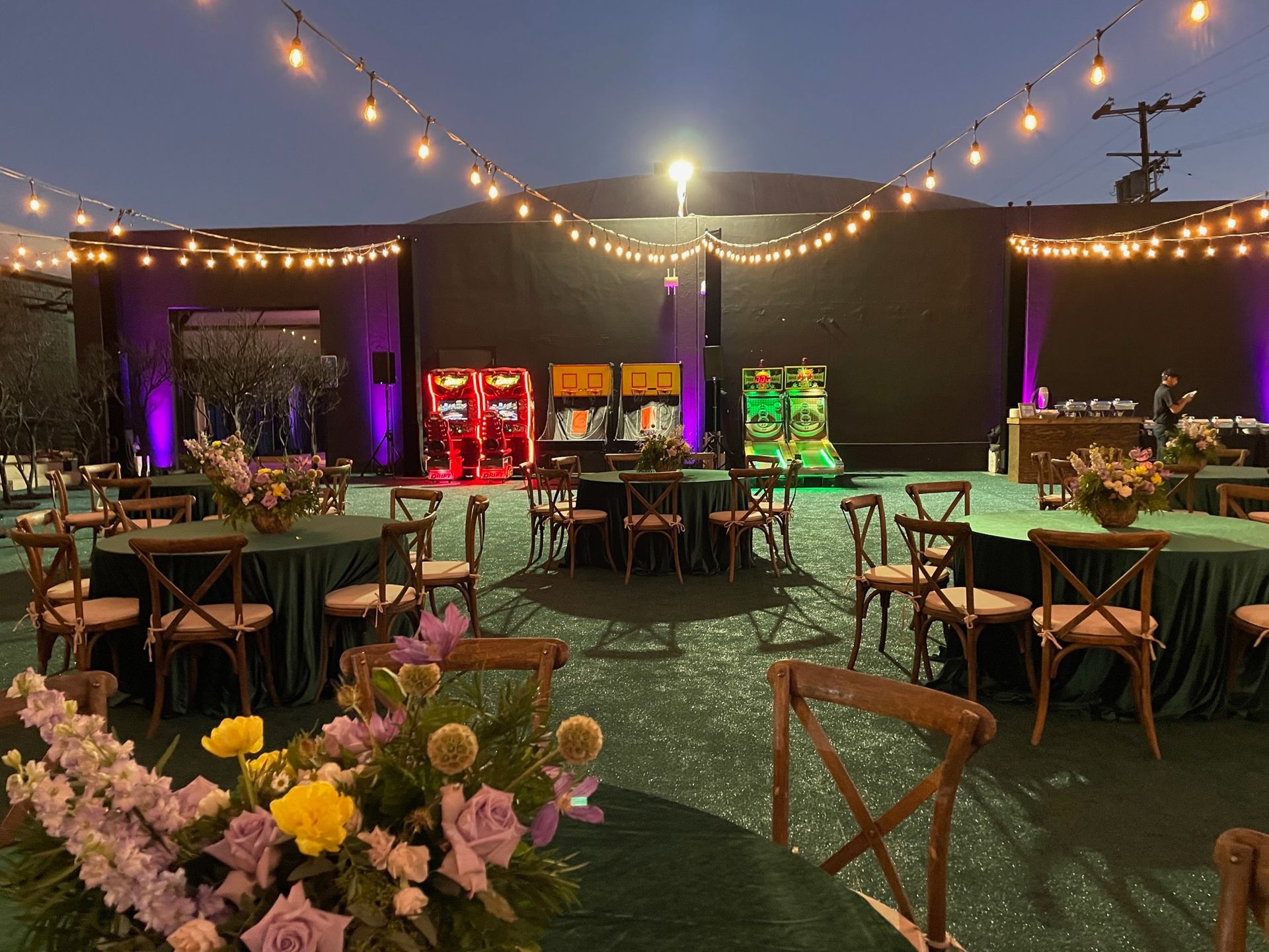 An outdoor evening event space with round tables, green tablecloths, floral centerpieces, and string lights overhead.