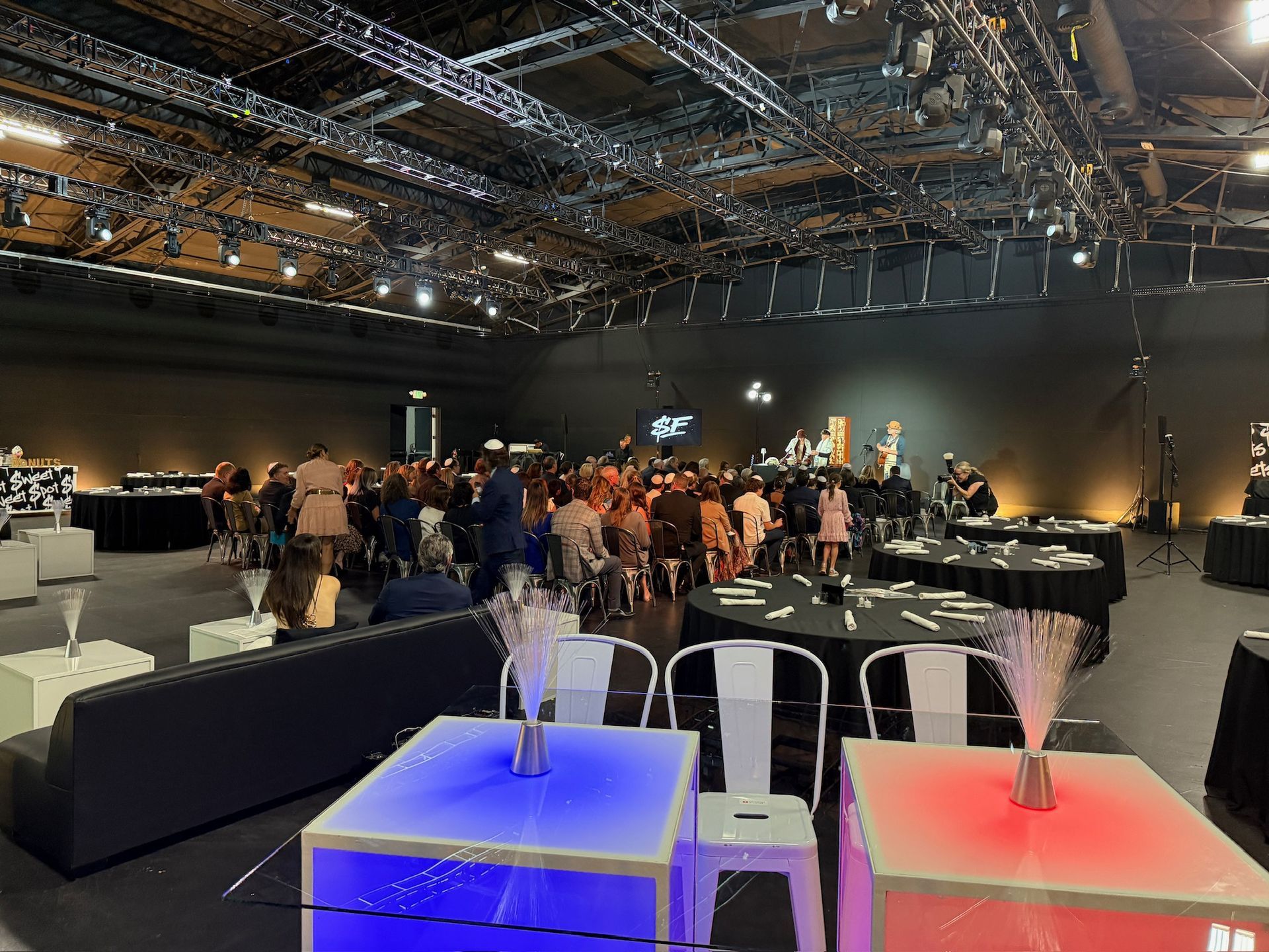 A dark, industrial event hall with lit-up tables in the foreground and a large group of people seated in the background.