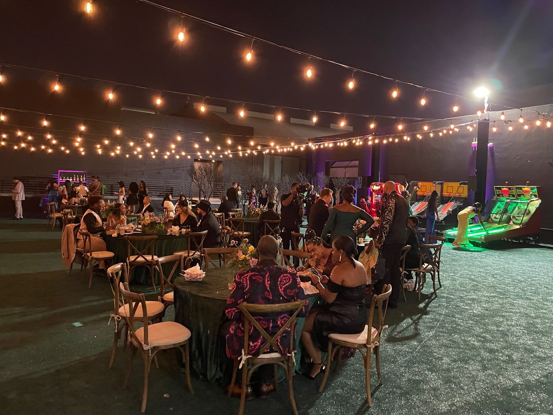 An outdoor evening event features guests seated at round tables under a canopy of string lights, with arcade games nearby.