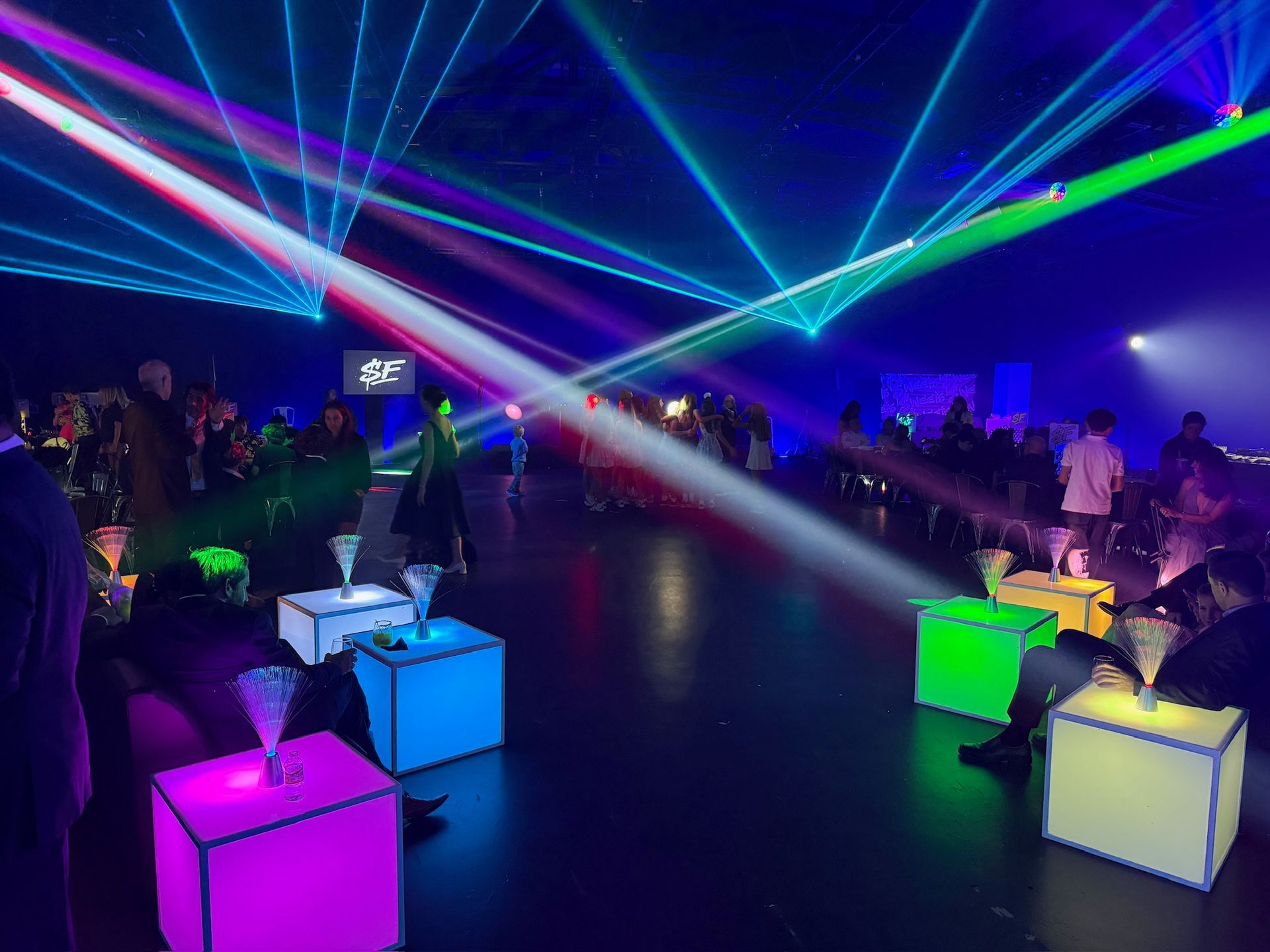 Event guests sitting on illuminated colorful cube stools under a display of vibrant laser lights in a dark room.