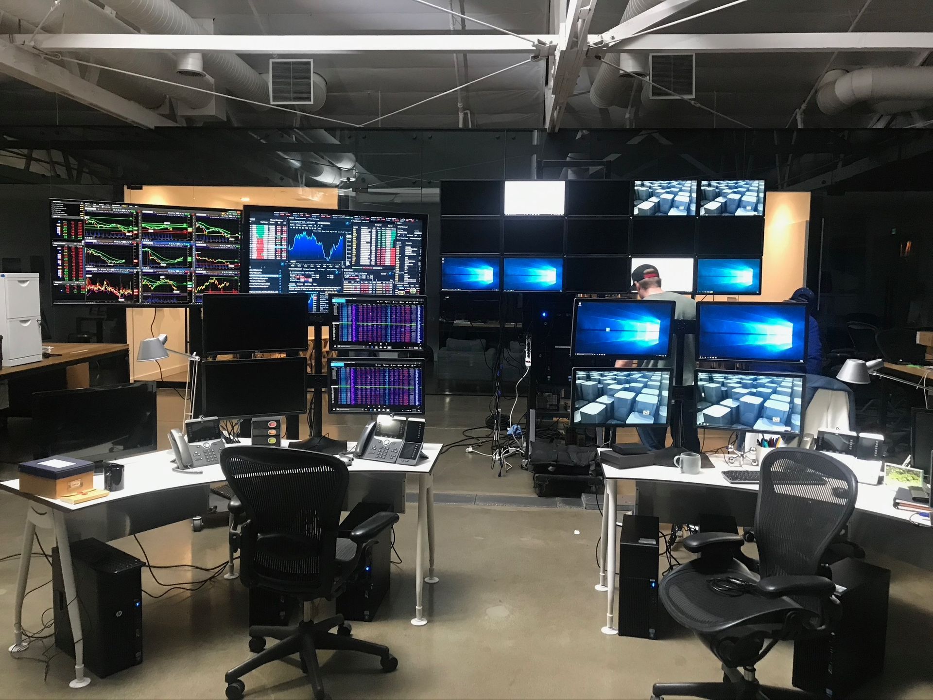 Two desk workstations in an office, each featuring multiple computer monitors displaying data charts and graphs.
