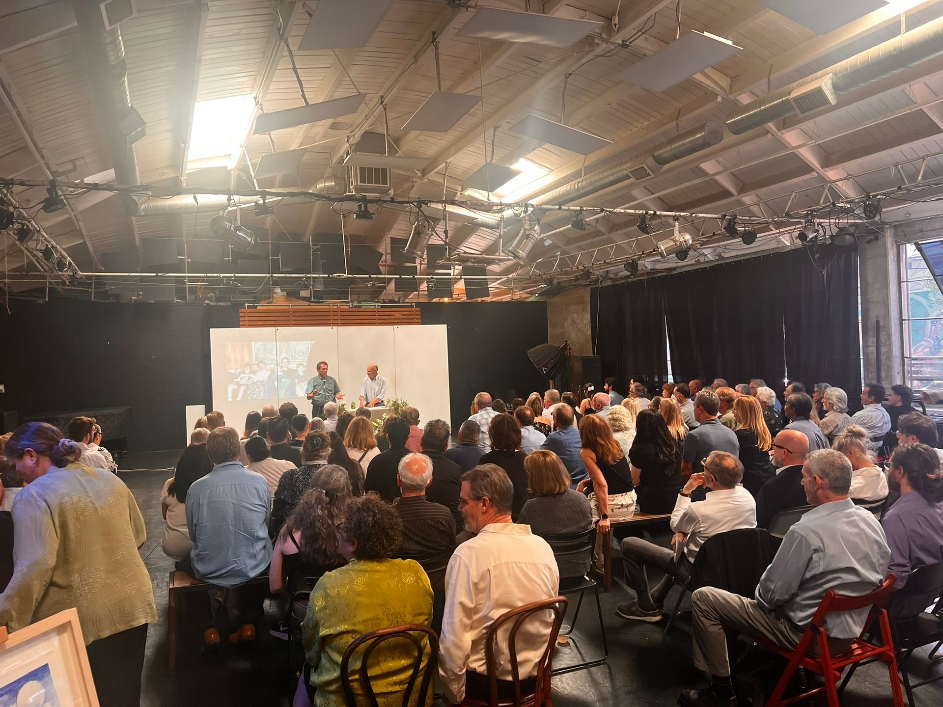 Audience seated in a large indoor venue watching two people present on a stage with a projection screen.
