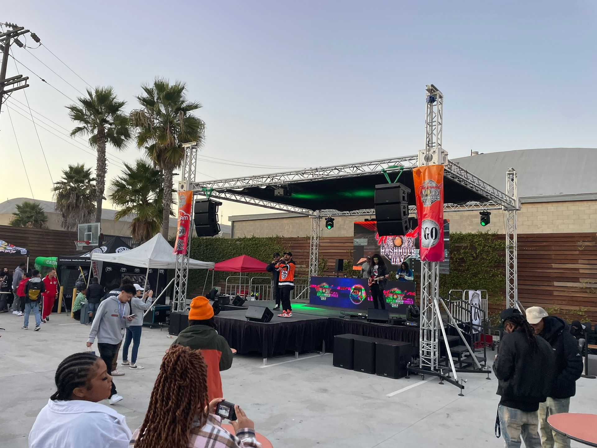 Performers on a stage at an outdoor event with a crowd gathered in a parking lot under a sunset sky.