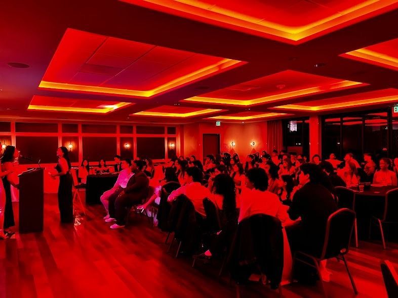 Two people speak at a podium before a seated audience in a dimly lit, red-toned event space with wooden floors.