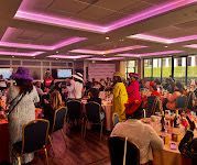 People in formal attire, some wearing hats, gather for a seated indoor event with pink recessed lighting.