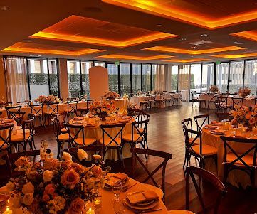 A formal dining room featuring round tables with floral centerpieces, wooden cross-back chairs, and warm recessed lighting.