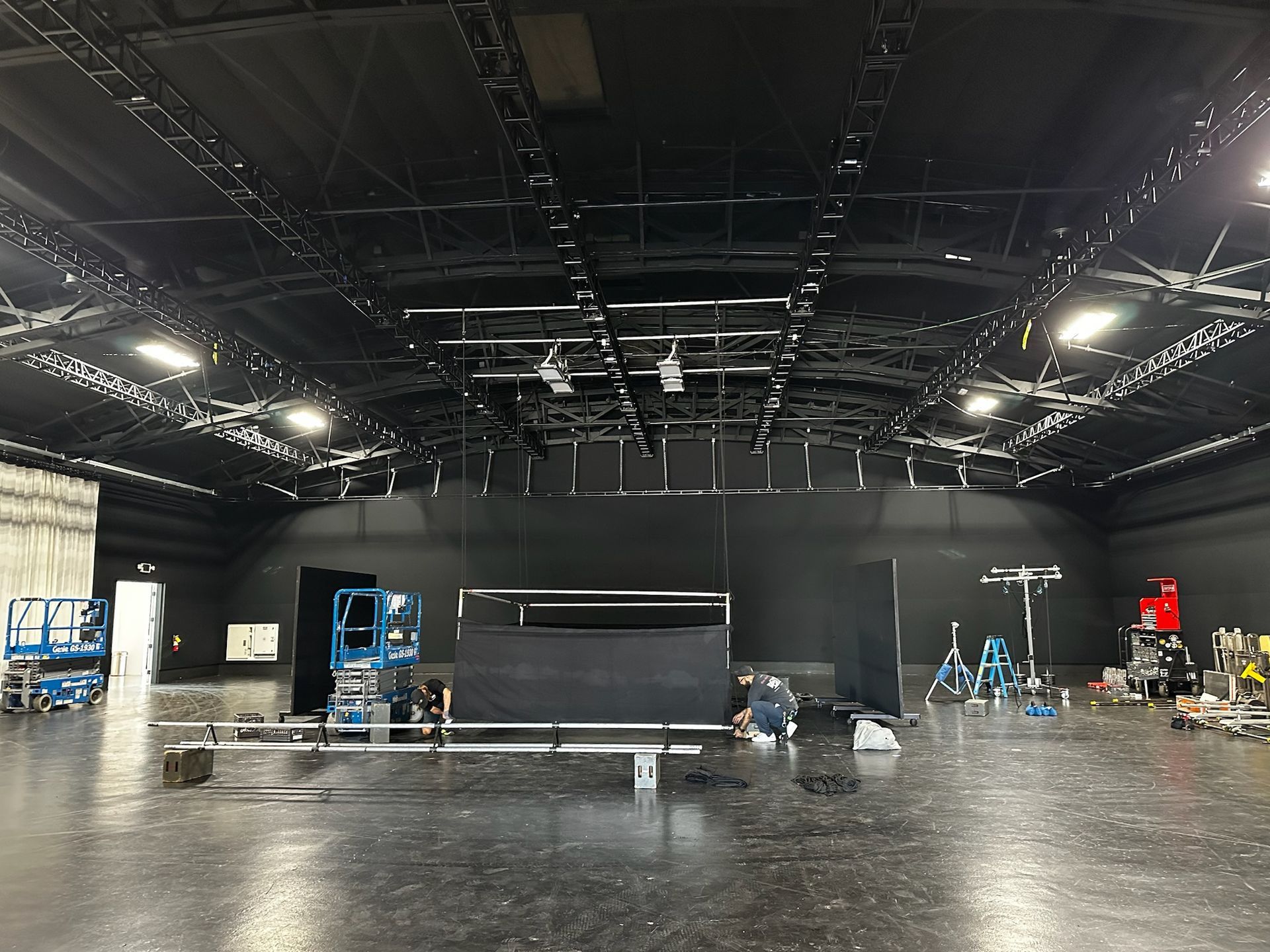 A large, empty film studio with black walls, a grid of overhead lights, and scattered production equipment on the floor.
