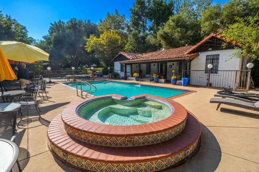 Outdoor pool with hot tub, lounge chairs, and a patio beside a white building on a sunny day