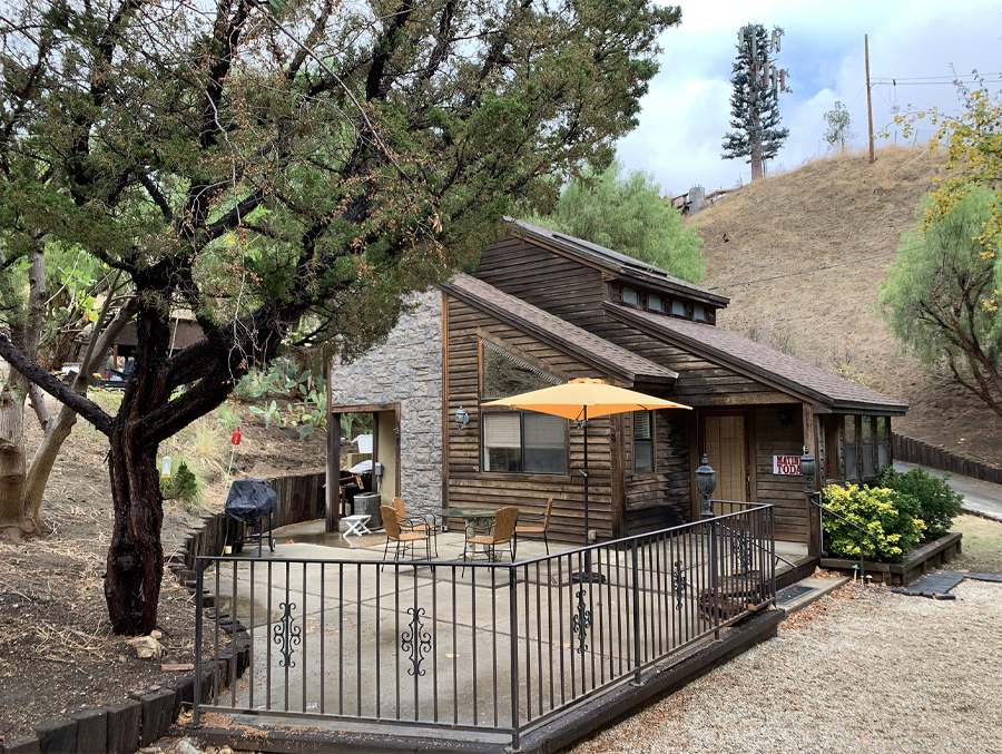 Stone cabin with sloped roof and patio railing on a wooded hillside, with a yellow umbrella outside
