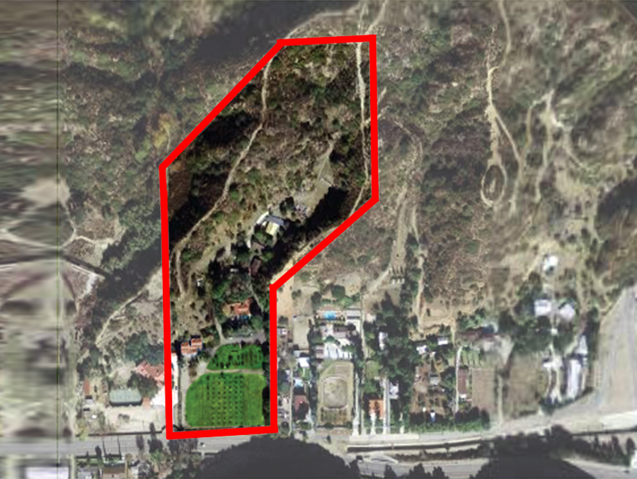 Aerial view of a wooded hillside outlined in red, beside buildings and a road.