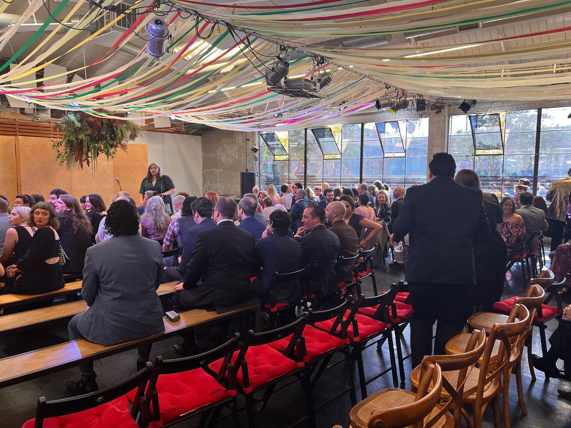 An indoor gathering features attendees seated in rows facing a stage under a ceiling decorated with hanging streamers.