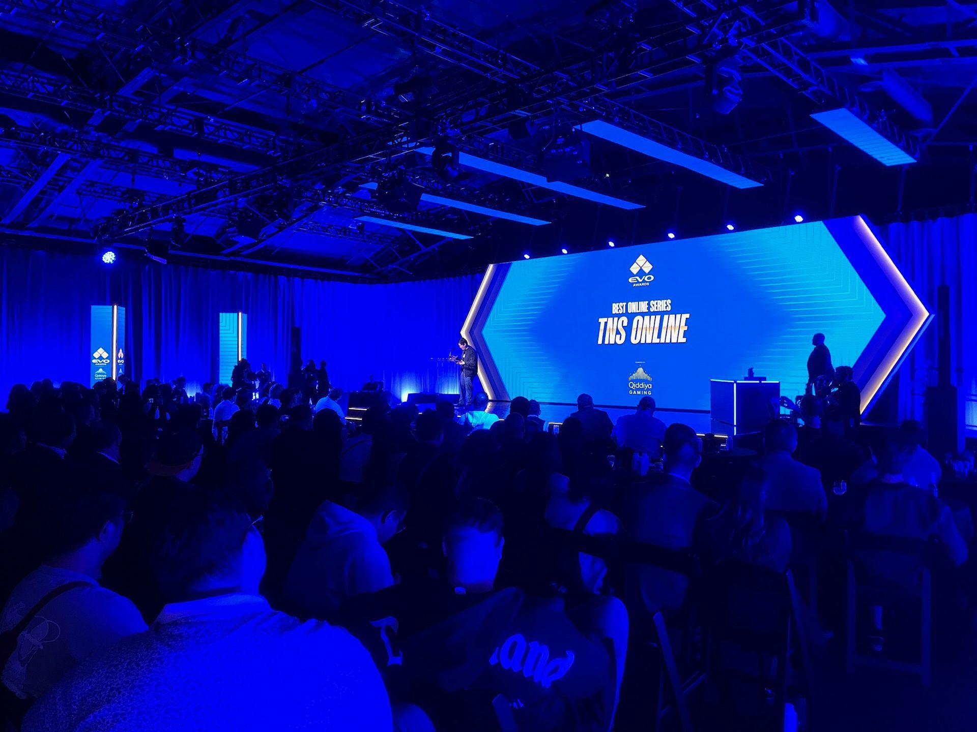 A large audience sits in a dimly lit, blue-toned room facing a stage featuring a glowing screen with event text.