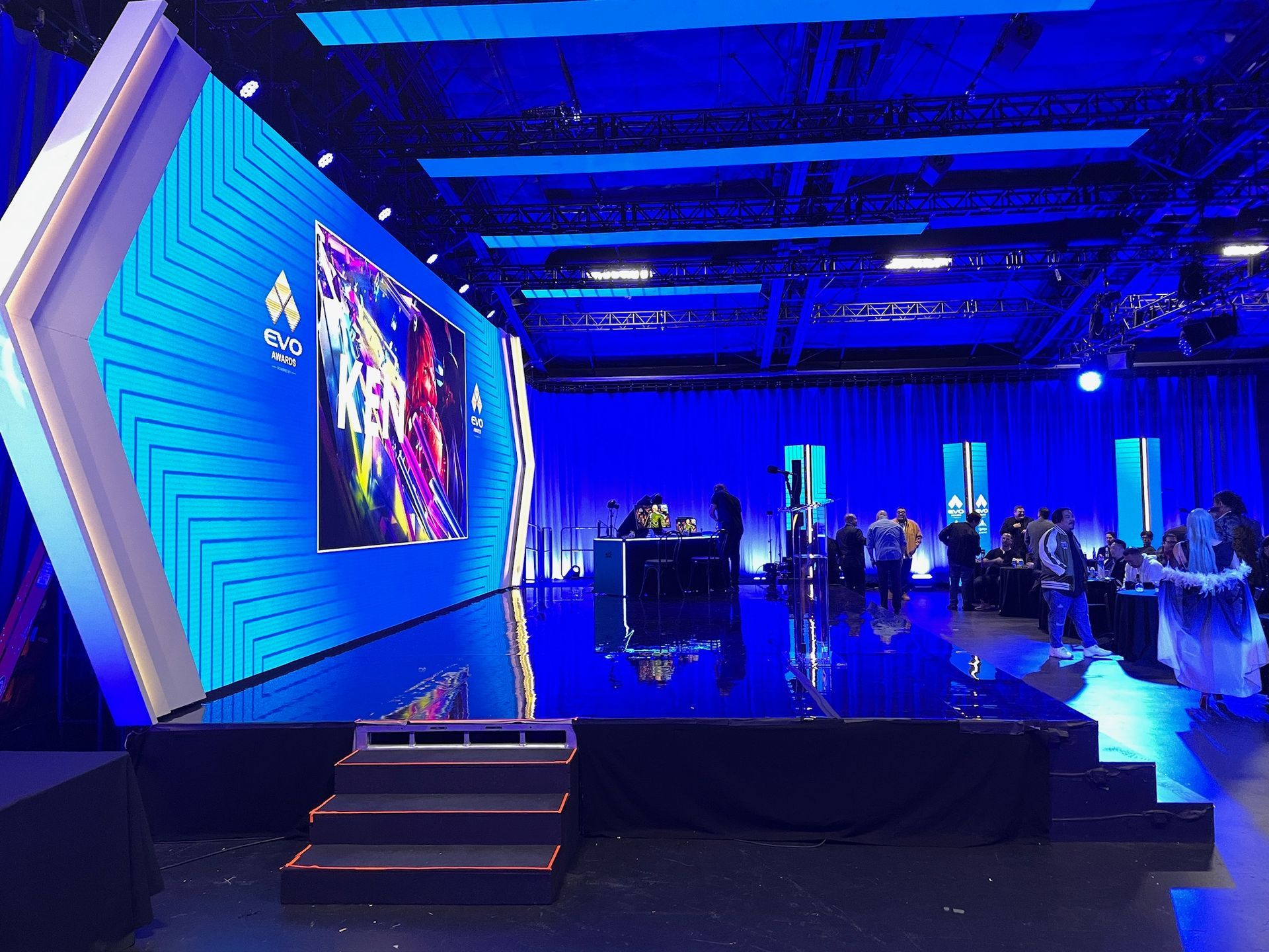 A stage with a large glowing blue screen and geometric accents in a dimly lit event space, with silhouettes of people.