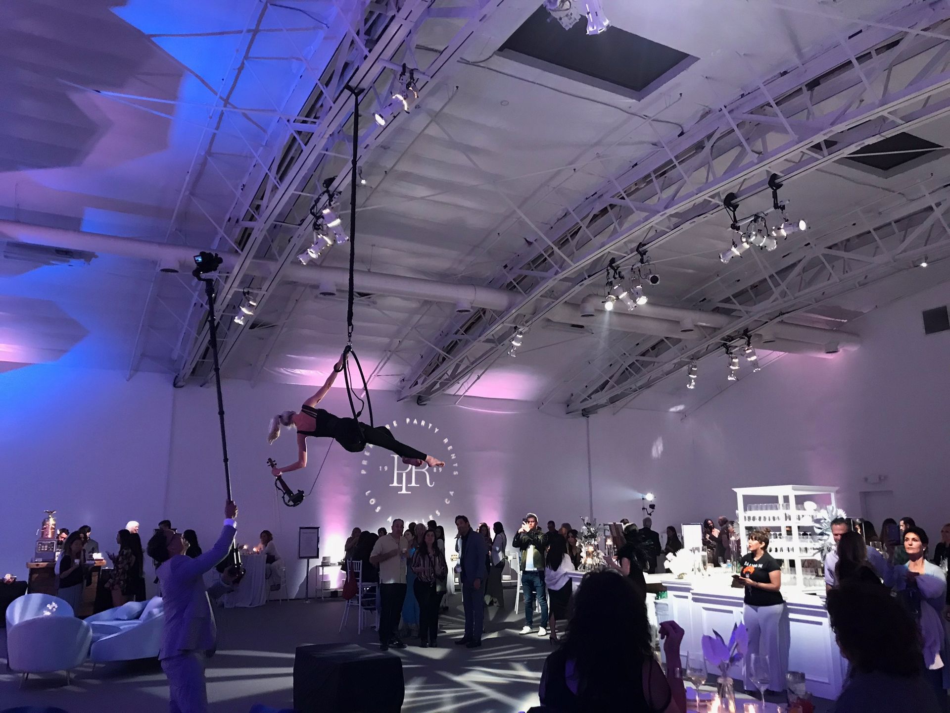 An aerial performer suspended from the ceiling entertains guests at a brightly lit, modern event space.