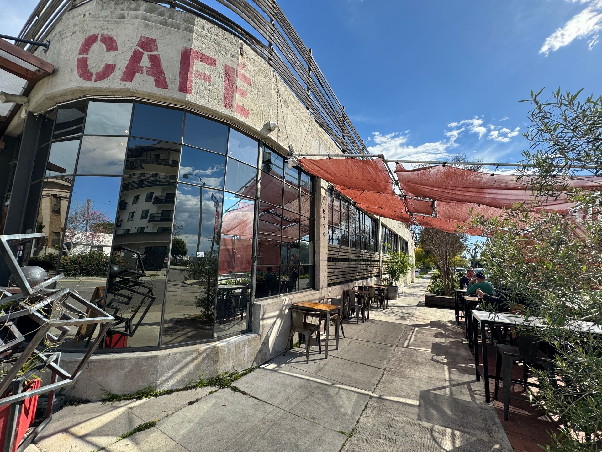 Outdoor patio seating at a cafe with reflective glass windows and a red shade canopy under a sunny blue sky.