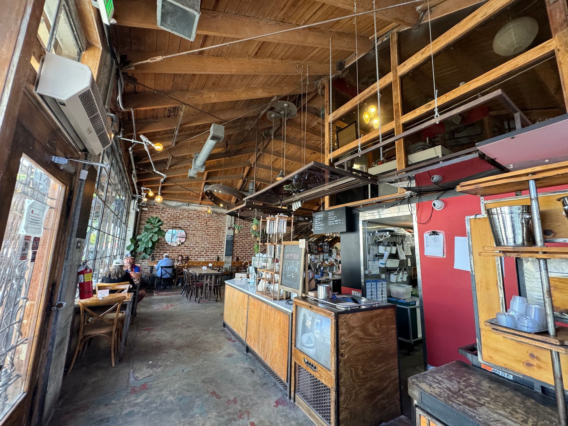 Interior of a rustic cafe with wood-beam ceilings, a brick wall, a service counter, and casual seating near glass windows.