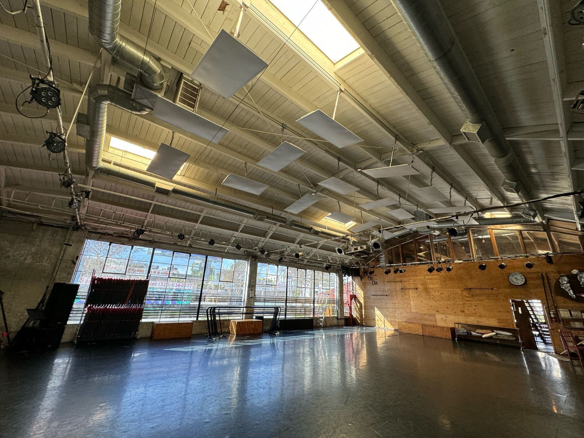 An empty, wide indoor dance studio with wooden walls, large windows, a reflective floor, and hanging acoustic panels.