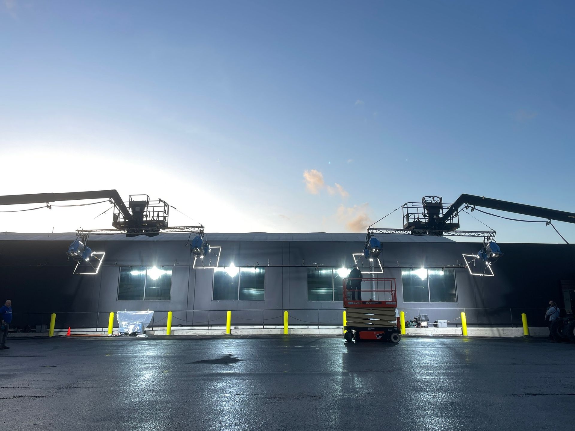 Two large boom lifts with mounted lights illuminate the exterior of a dark commercial building under a clear blue sky.