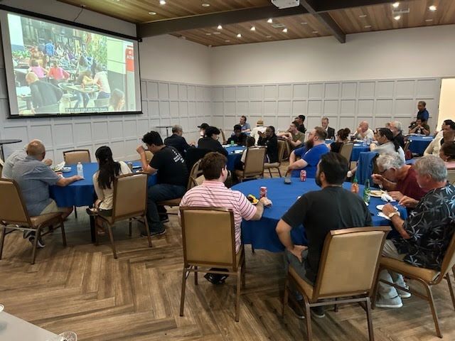 A group of people are sitting at tables in front of a large screen.