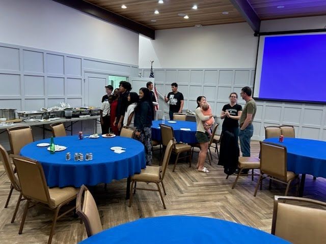 A group of people are standing around tables in a room with blue tables and chairs.
