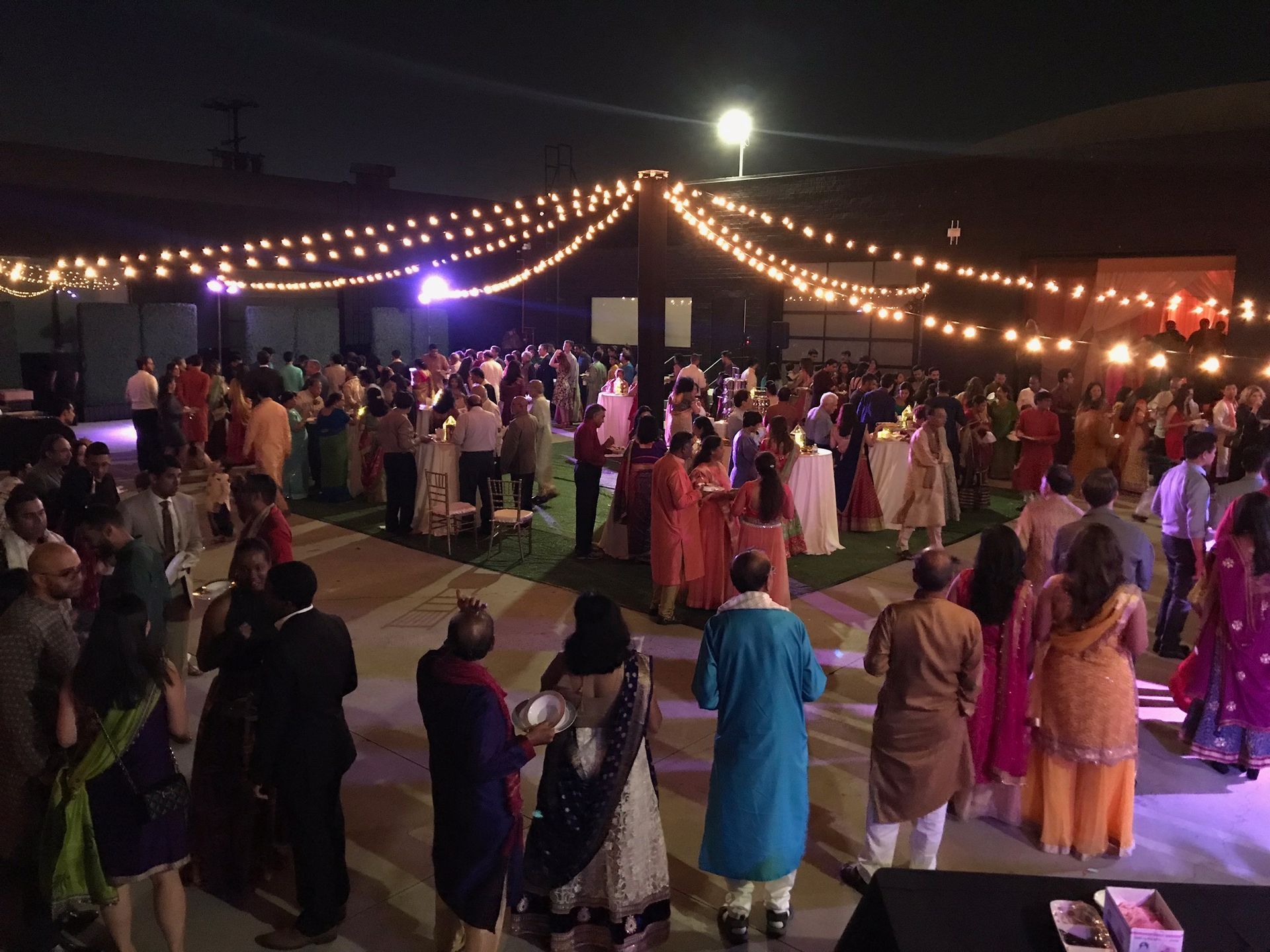 Guests at an evening outdoor event mingle beneath strings of warm fairy lights on a paved patio.