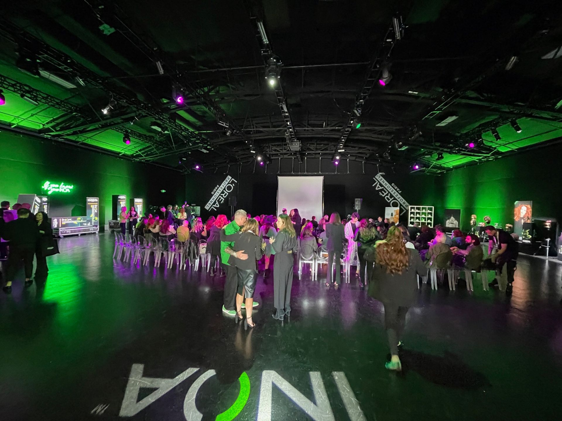 A dimly lit, green-hued event hall with attendees mingling in front of seated rows and a large screen.