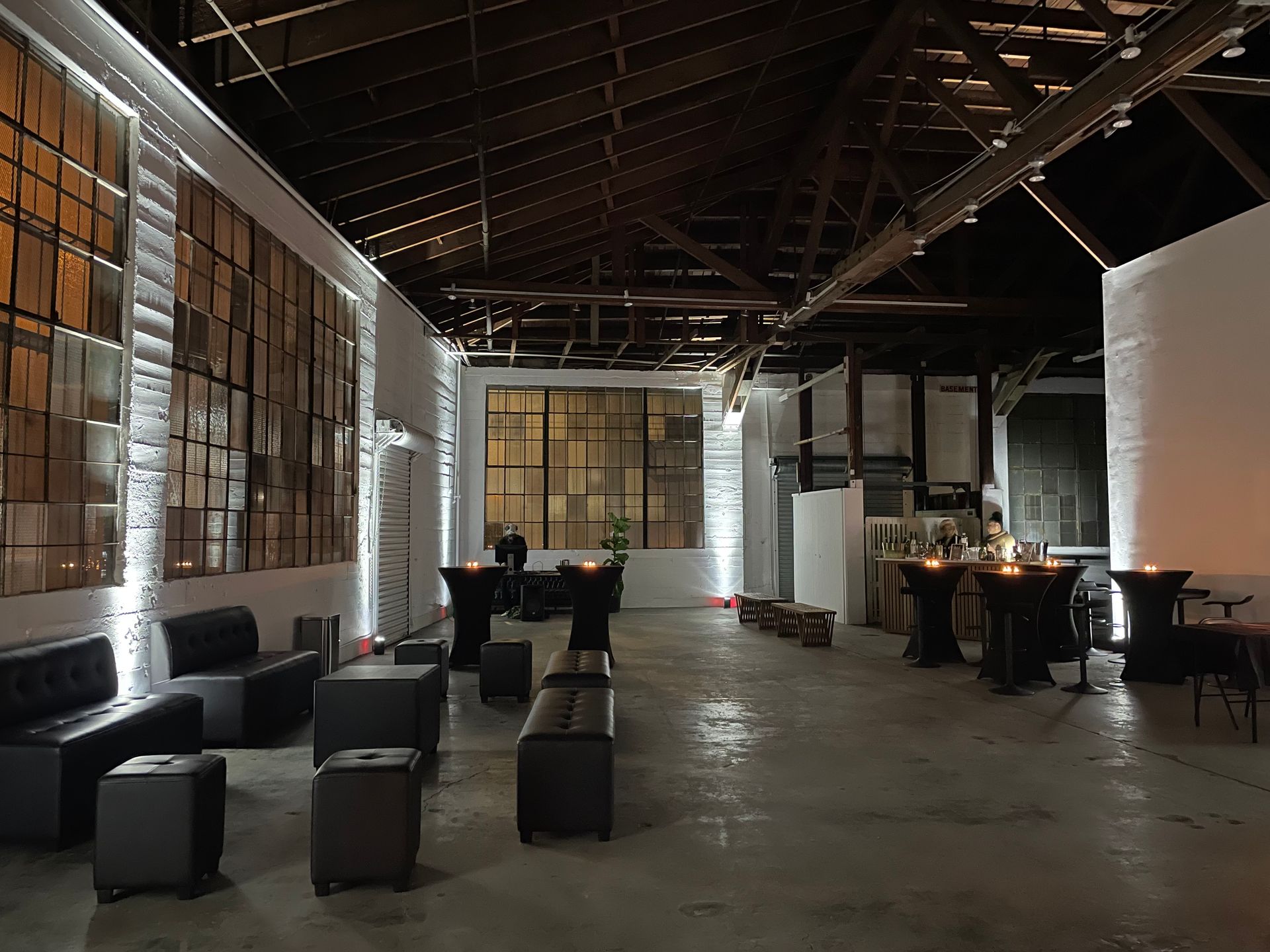 An open, industrial event space with dark modern furniture, high timber ceilings, and large grid windows.