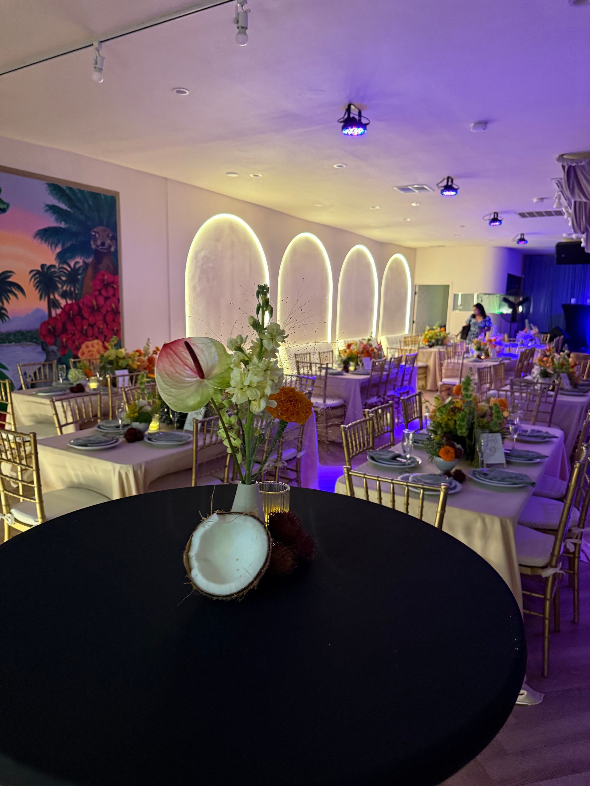 Banquet hall with round tables, floral centerpieces, purple lighting, and illuminated wall arches