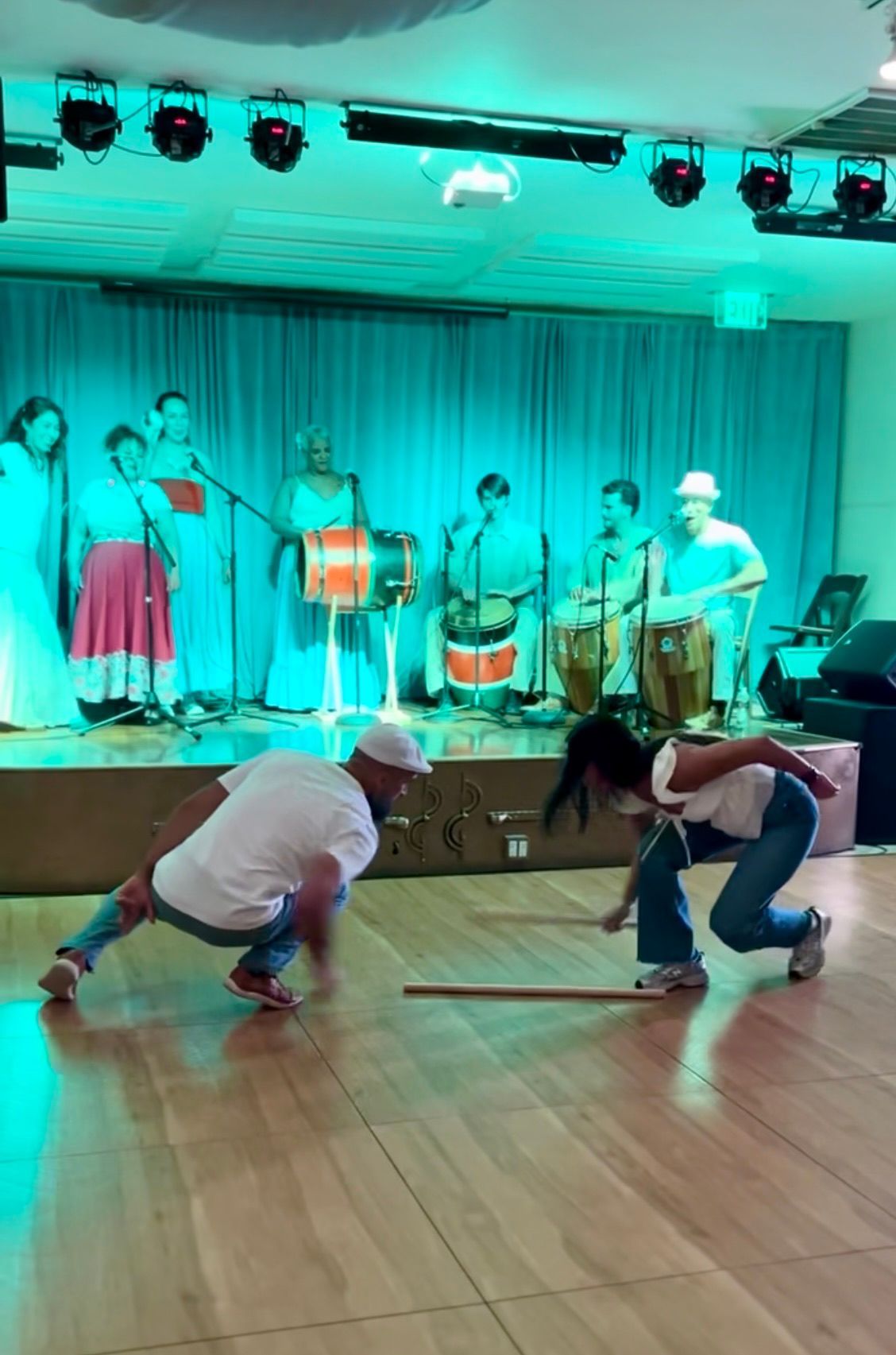 Two people crouch on a dance floor before a lit stage with musicians and teal curtains