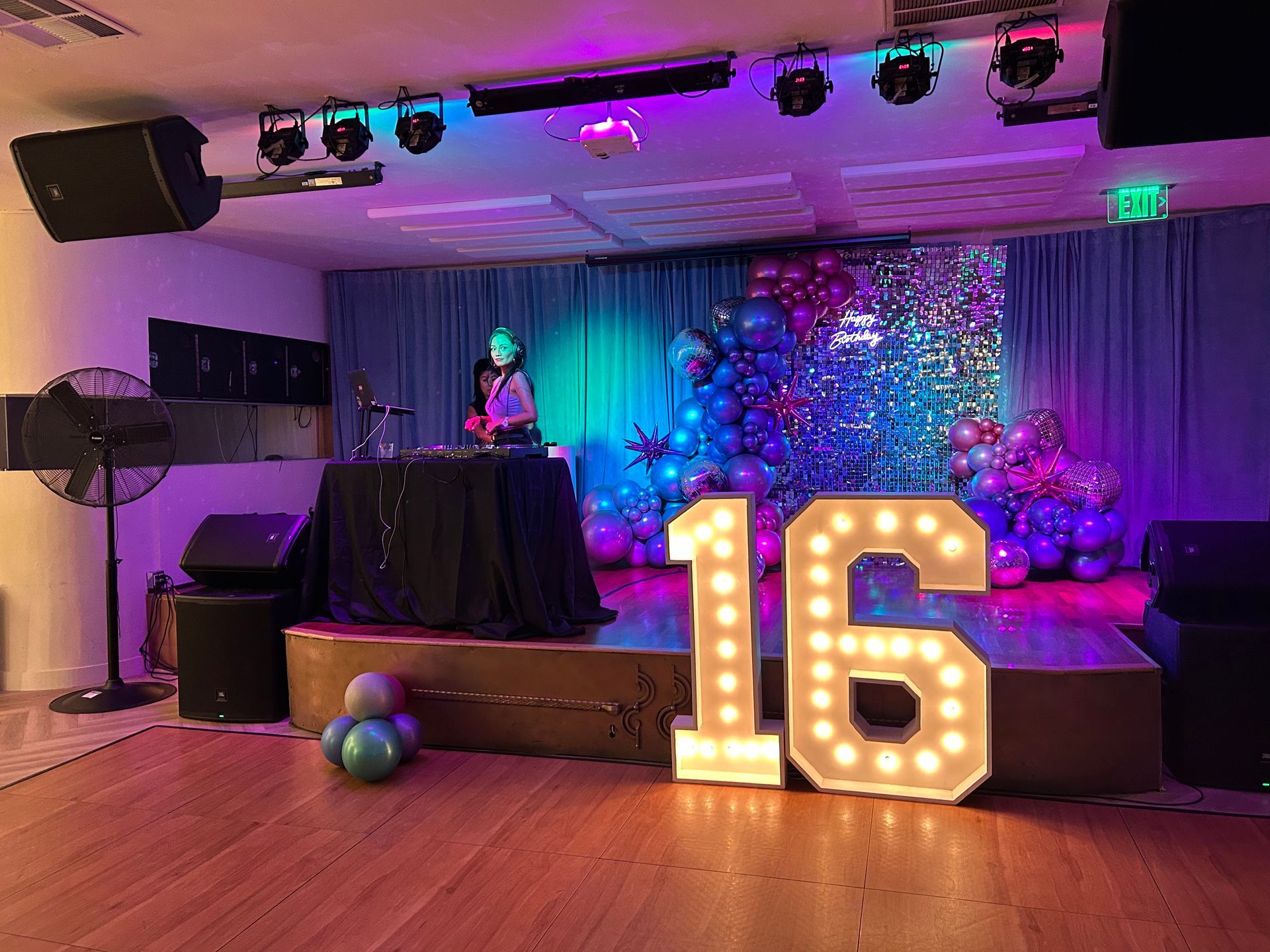 Decorated party stage with a lit “16,” DJ booth, colorful lights, and balloon decorations